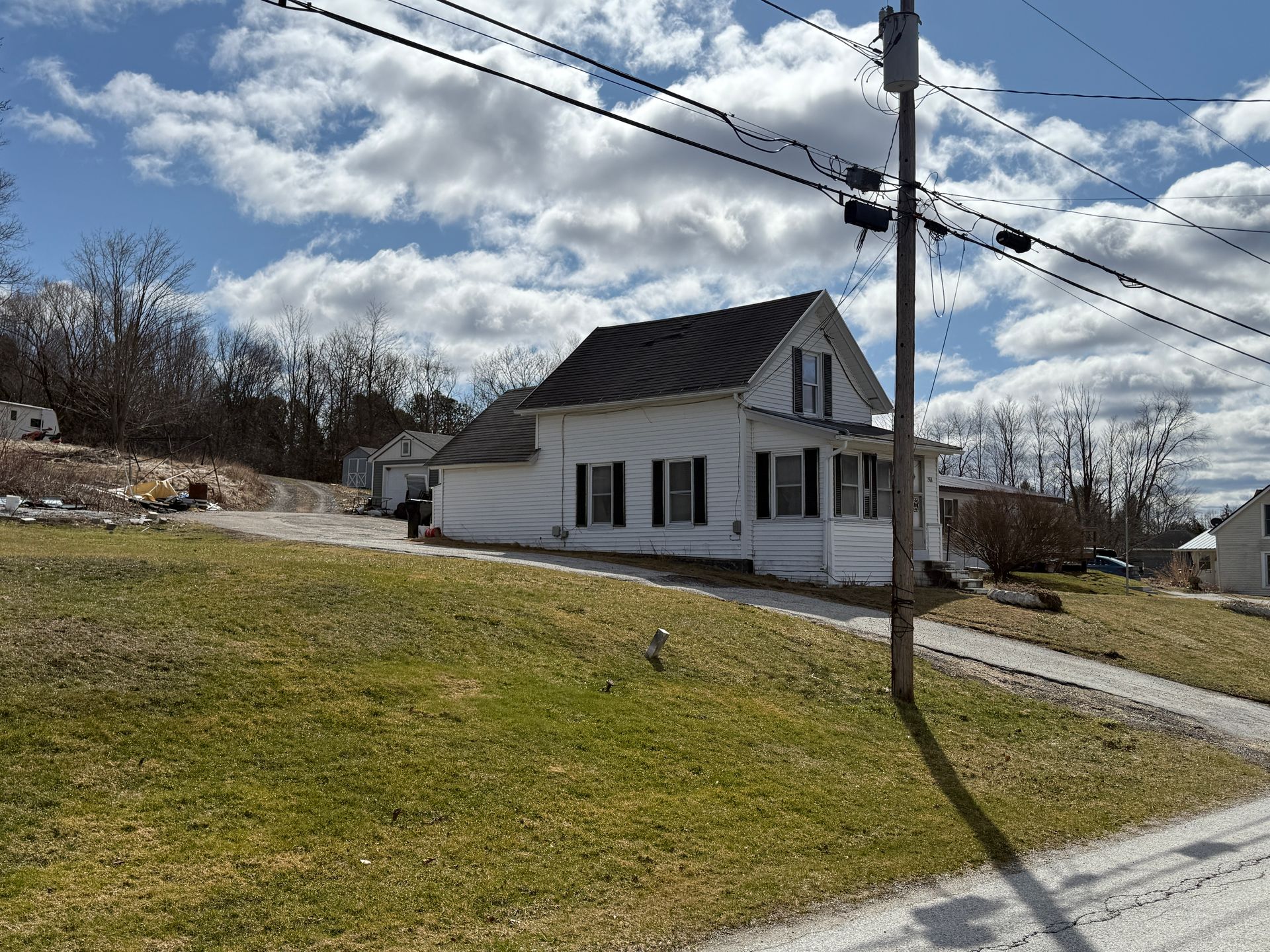 White house with black shutters on a grassy hill under a partly cloudy sky.