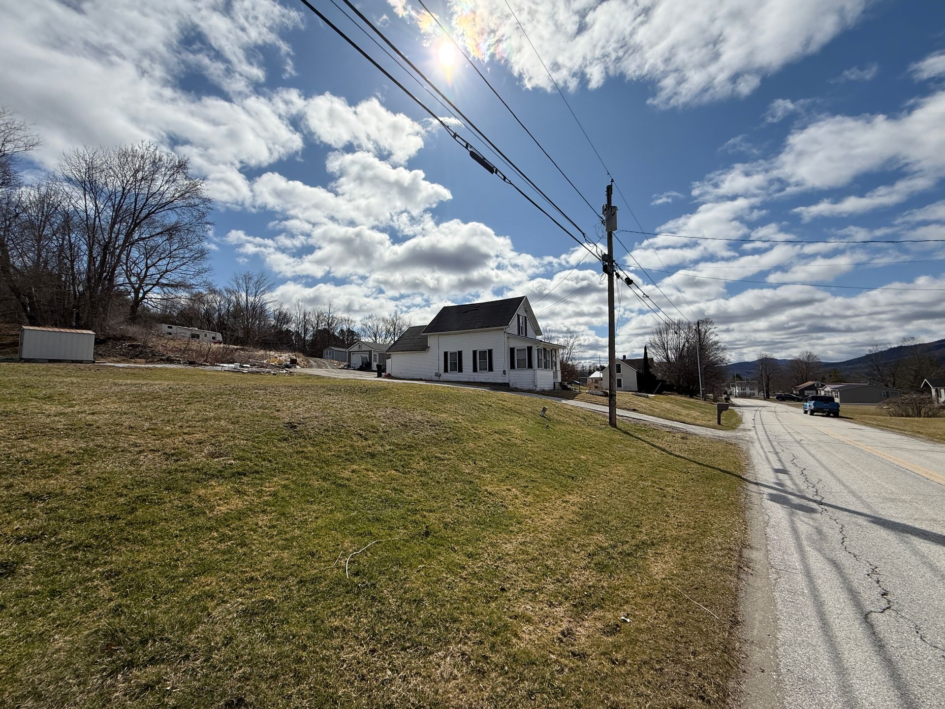 White house on a grassy hill under a bright sun and cloudy sky. Power lines and a road in the foreground.