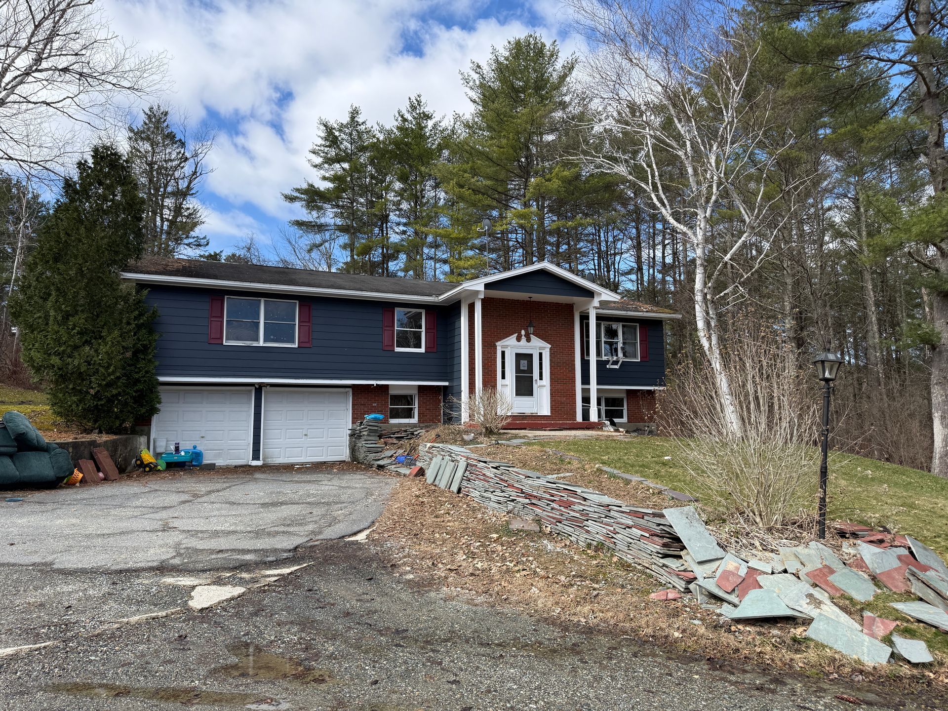 House with a blue exterior and brick entrance. Driveway, stone retaining wall, and trees in background.