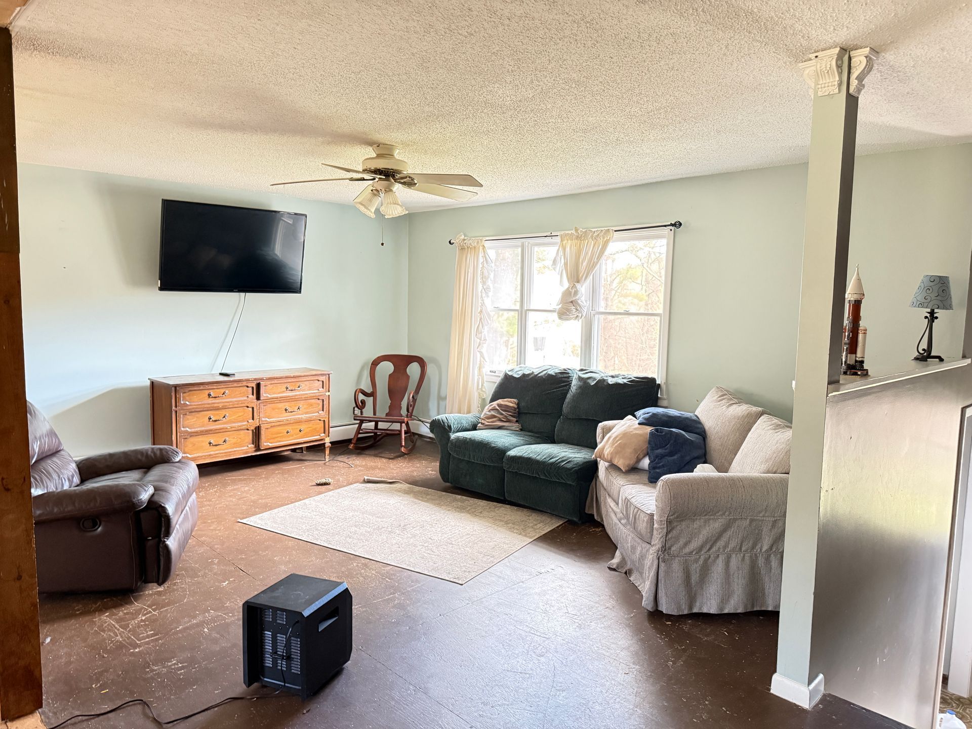 Living room with couch, TV, dresser, and rug on a dark floor with a window and a ceiling fan.