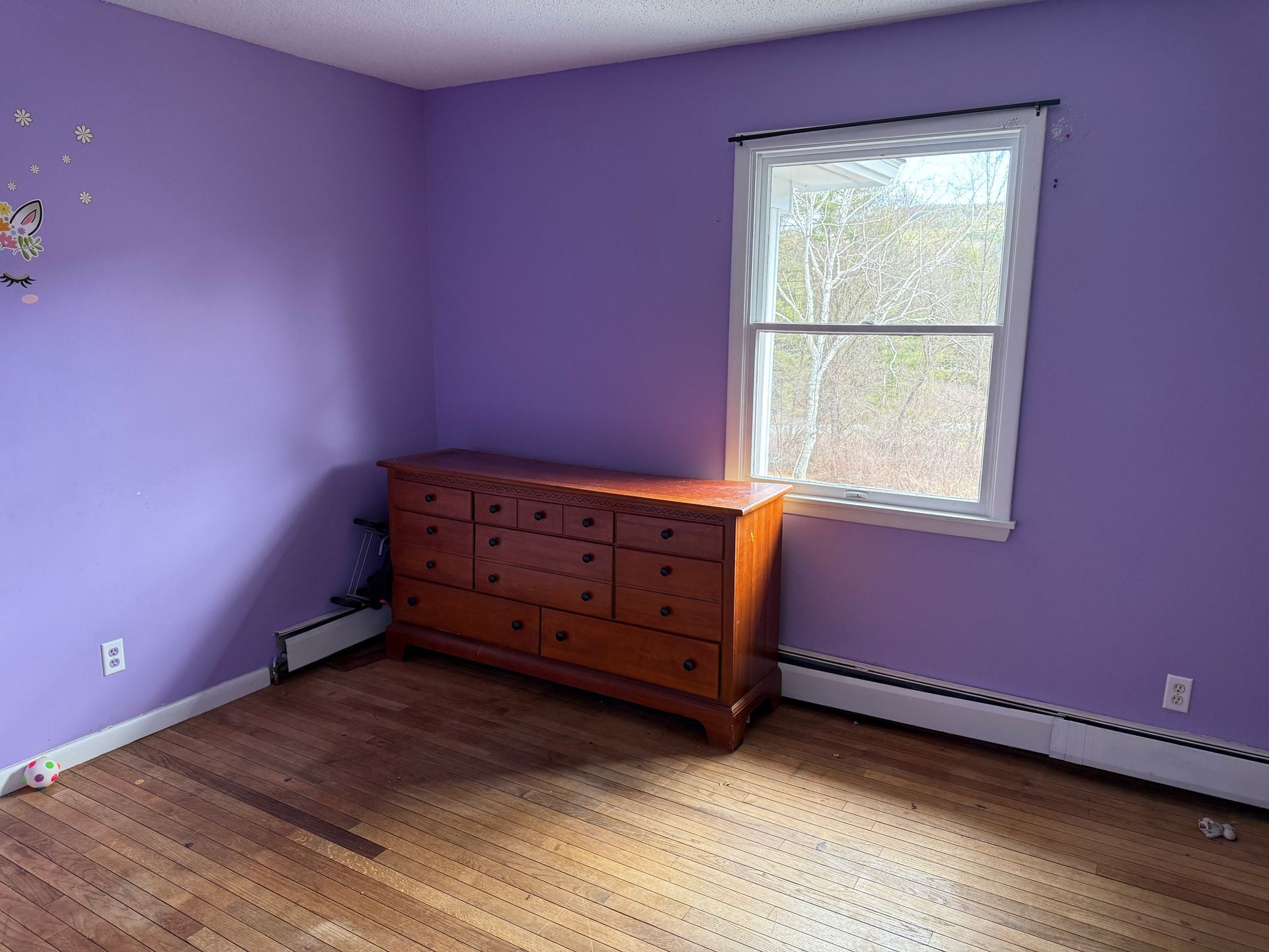 Bedroom with purple walls, wooden dresser, window, and hardwood floor.