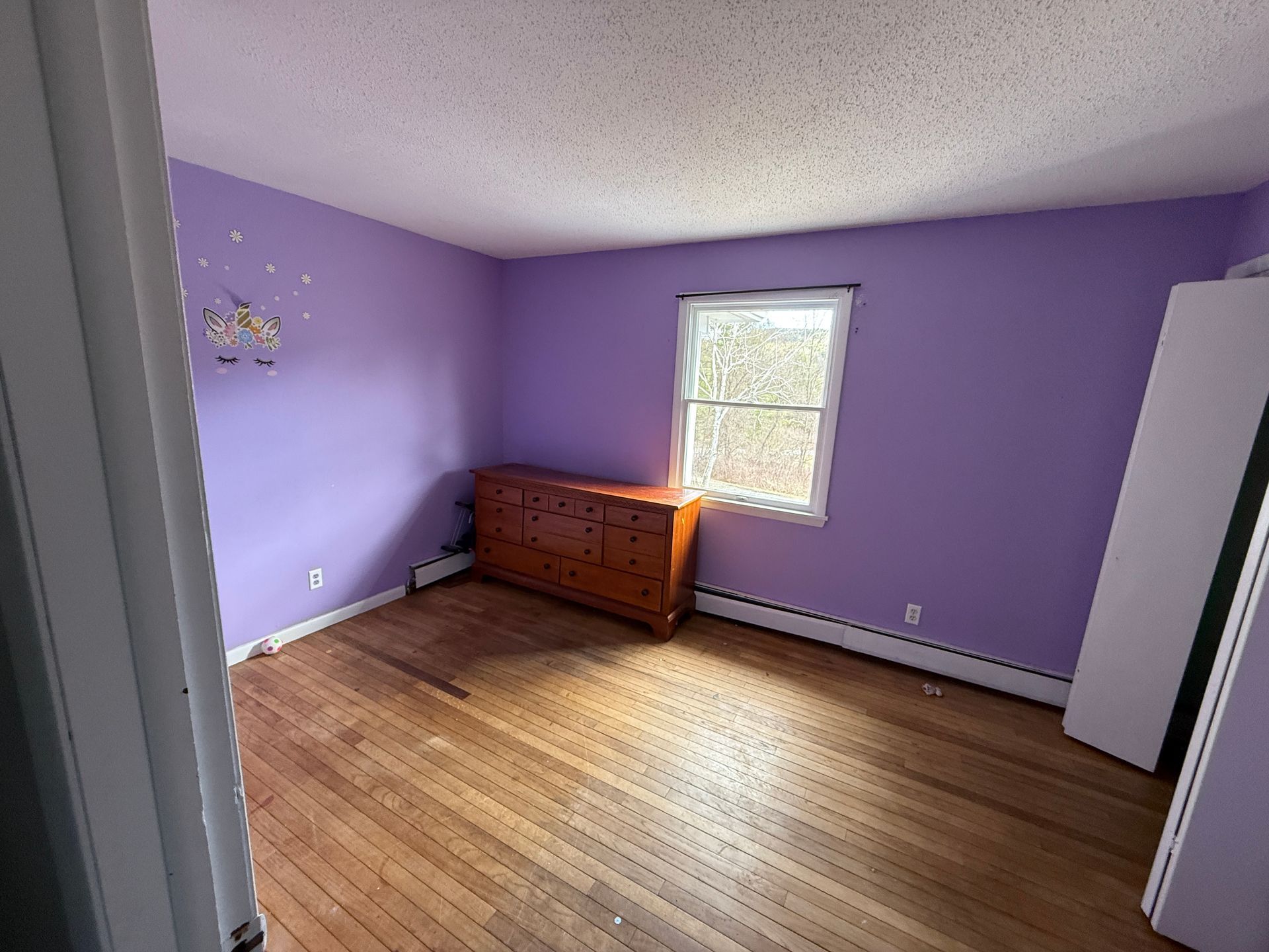 A room with purple walls, wooden floor, window, and a brown dresser.