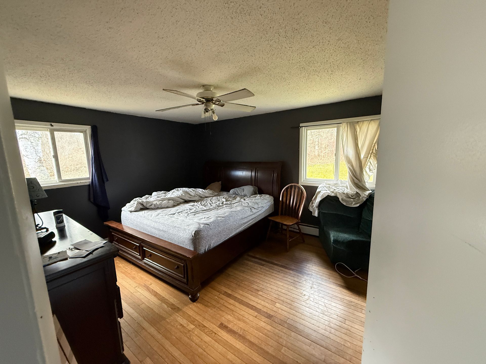 Bedroom with dark walls, wooden bed, and windows. A chair and armchair are also present.