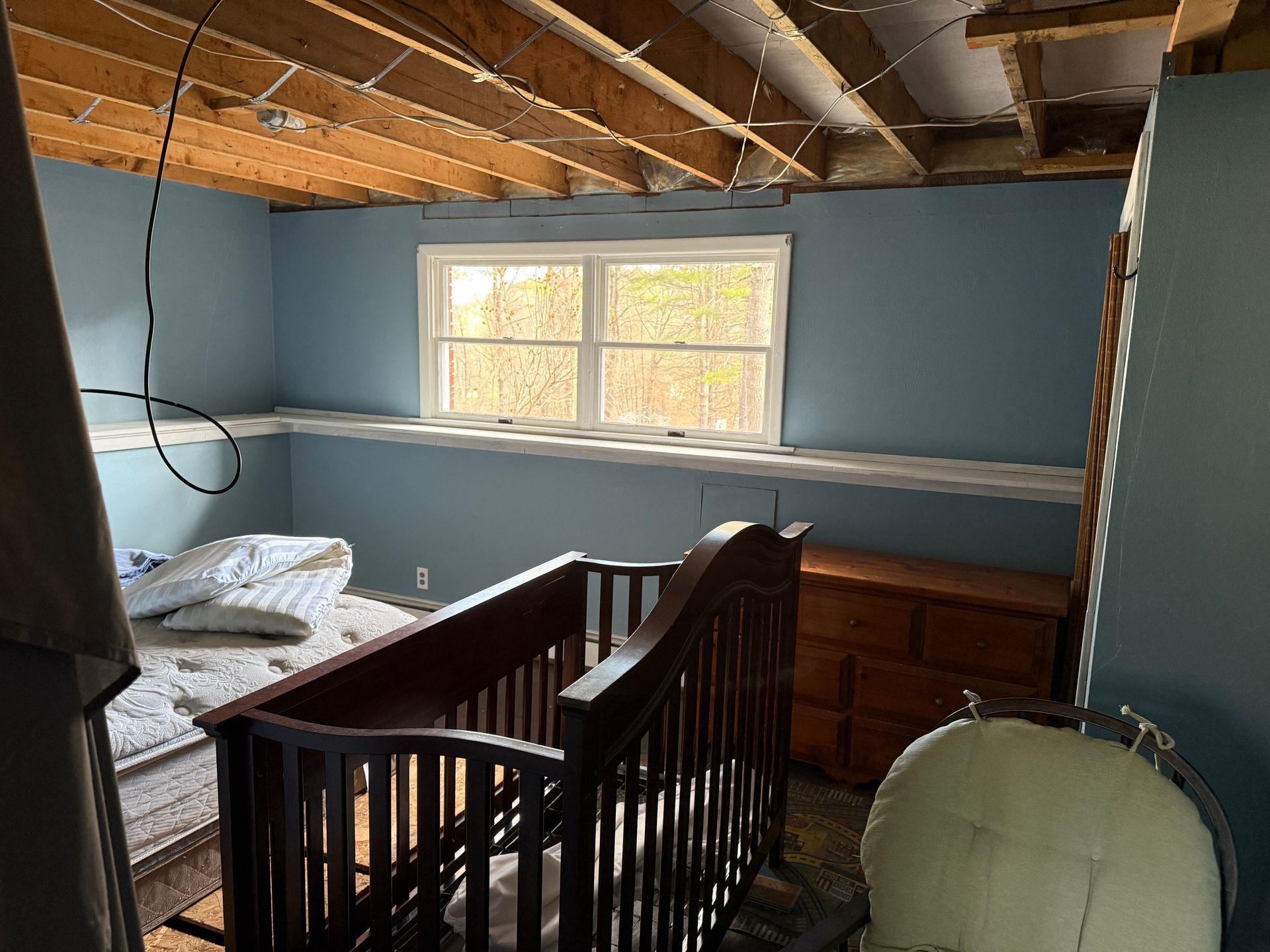 Basement room with blue walls, crib, chest of drawers, window, and exposed ceiling beams.