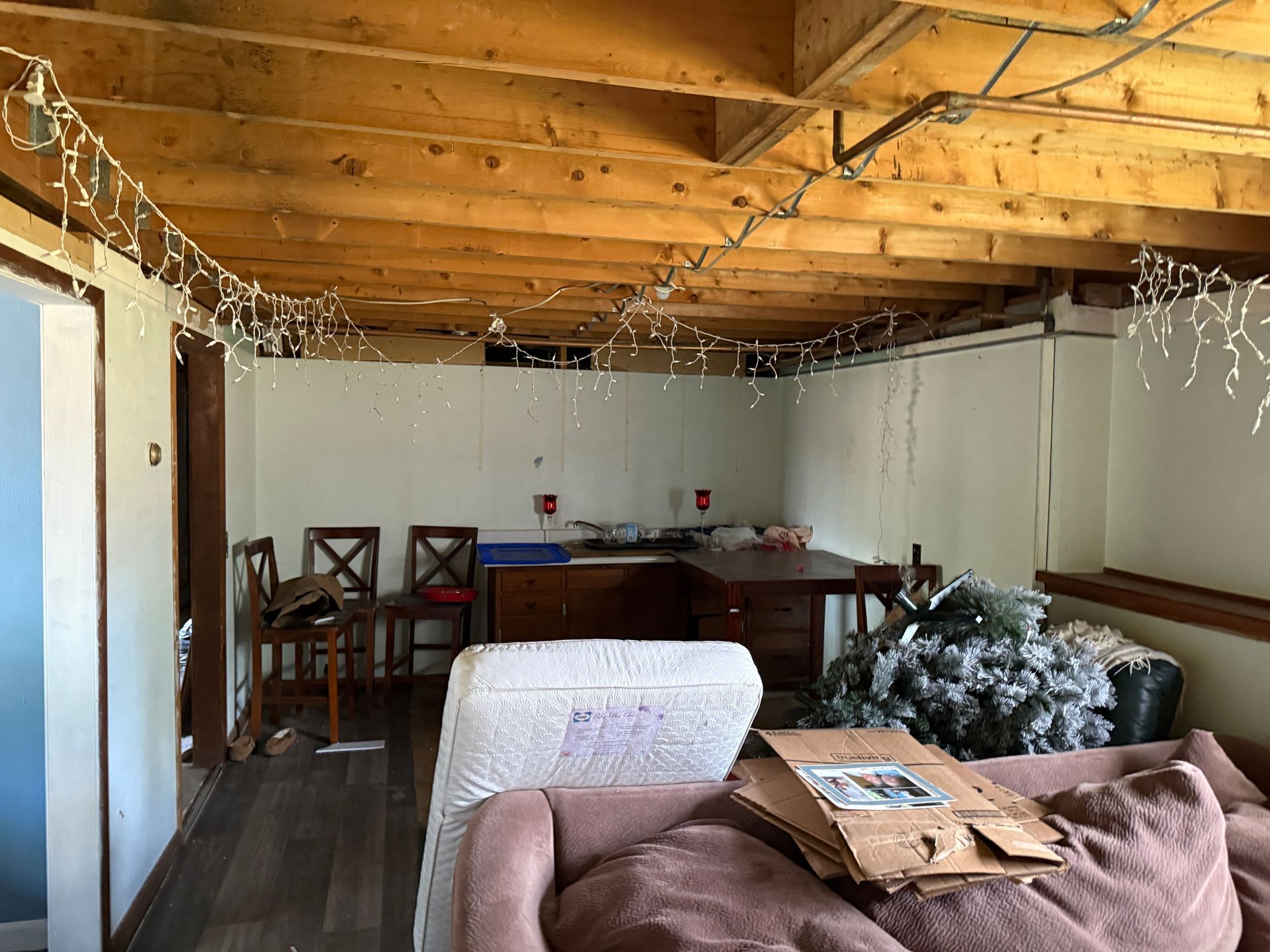 Basement interior with exposed wooden ceiling, white walls, furniture, and Christmas lights.