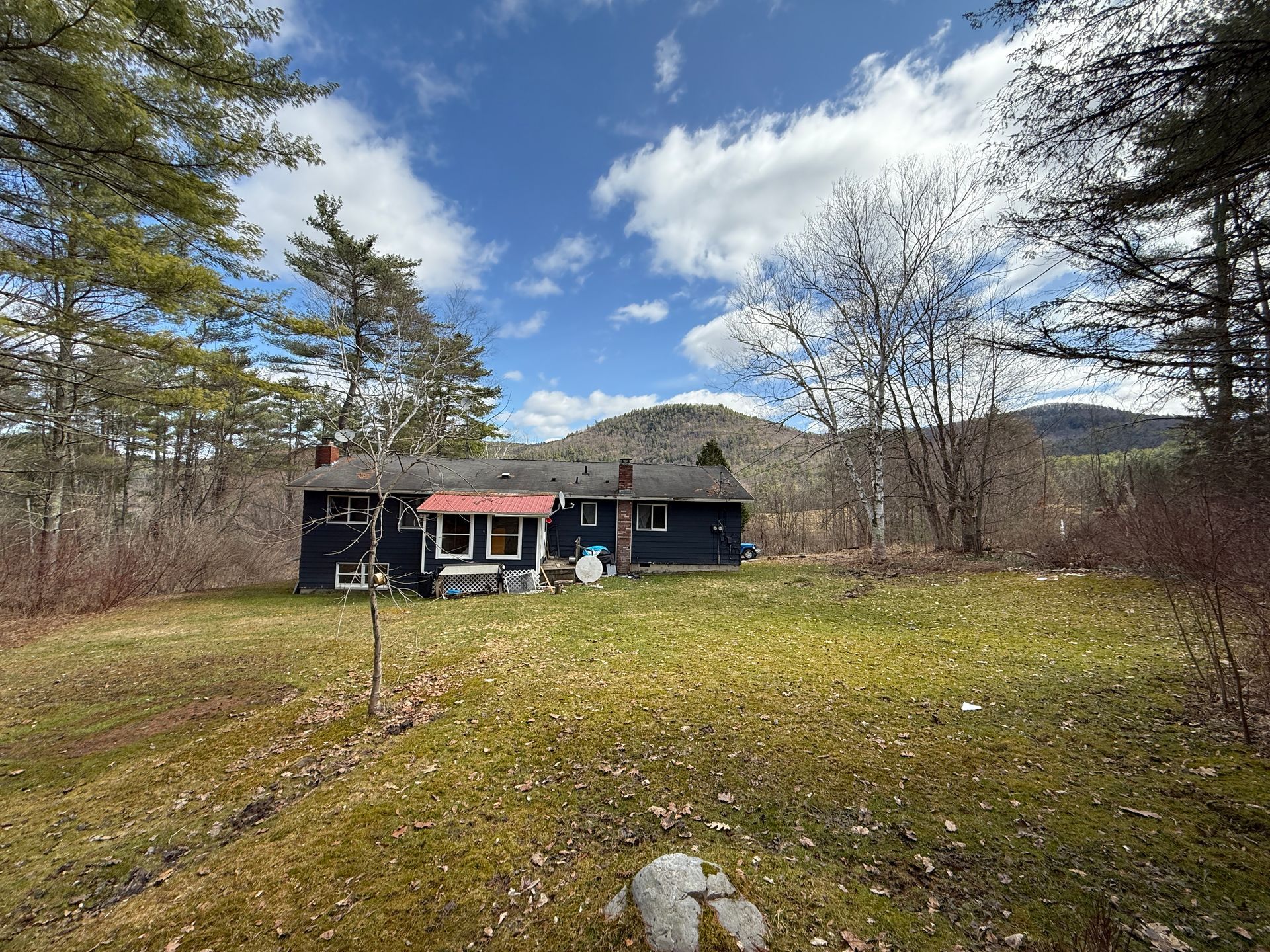 A dark house in a grassy yard, surrounded by trees under a partly cloudy sky.