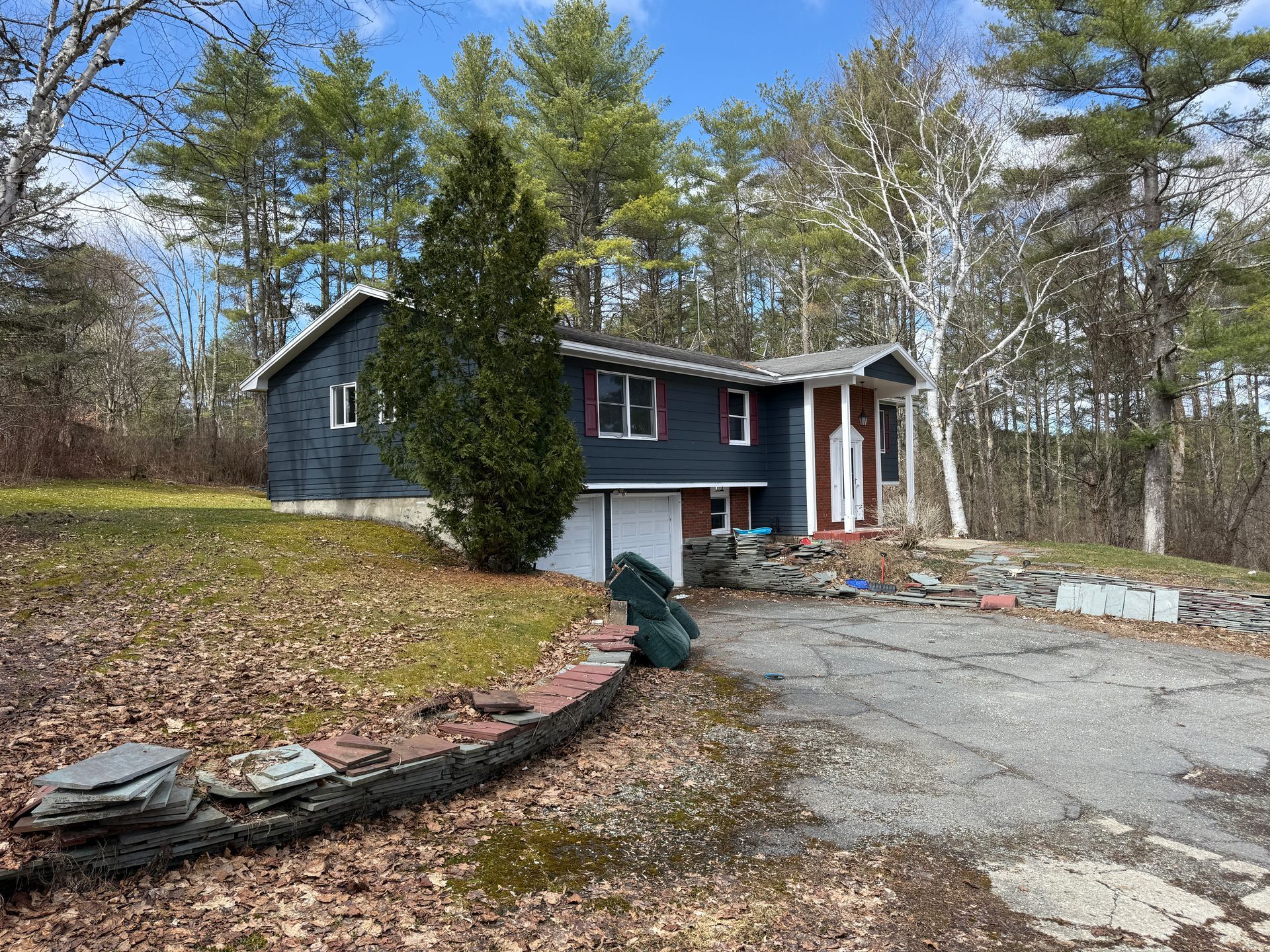 Dark-colored house with white trim, driveway, and autumn leaves; surrounded by trees under a partly cloudy sky.