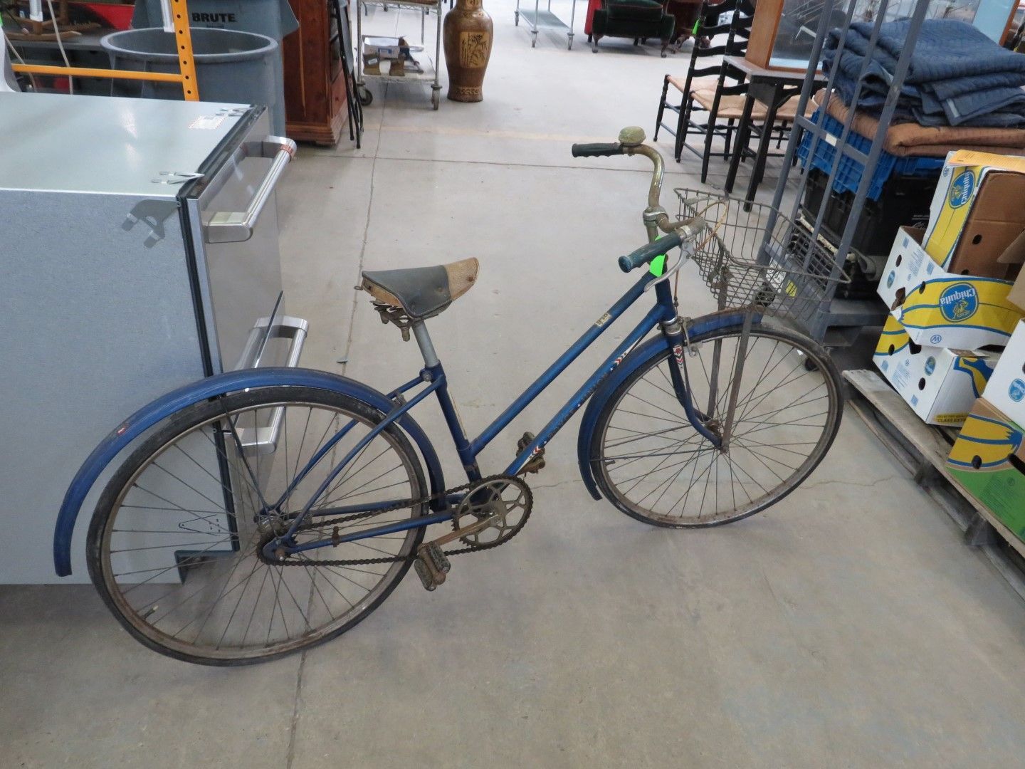 Blue vintage bicycle with a basket and mudguards on a concrete floor.
