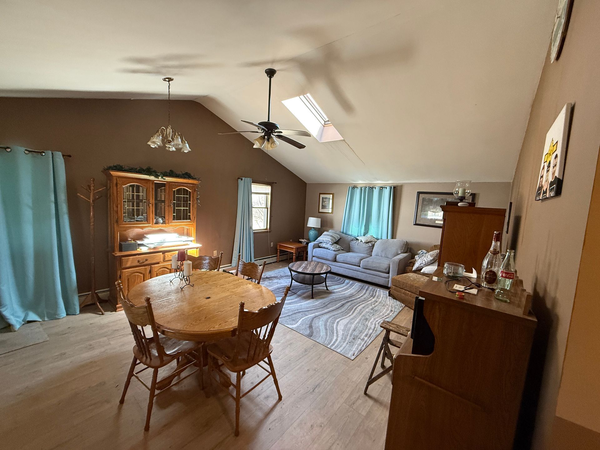 Living room with dining table, hutch, sofa, rug, and skylight. Light blue curtains, brown wall, light wood floor.