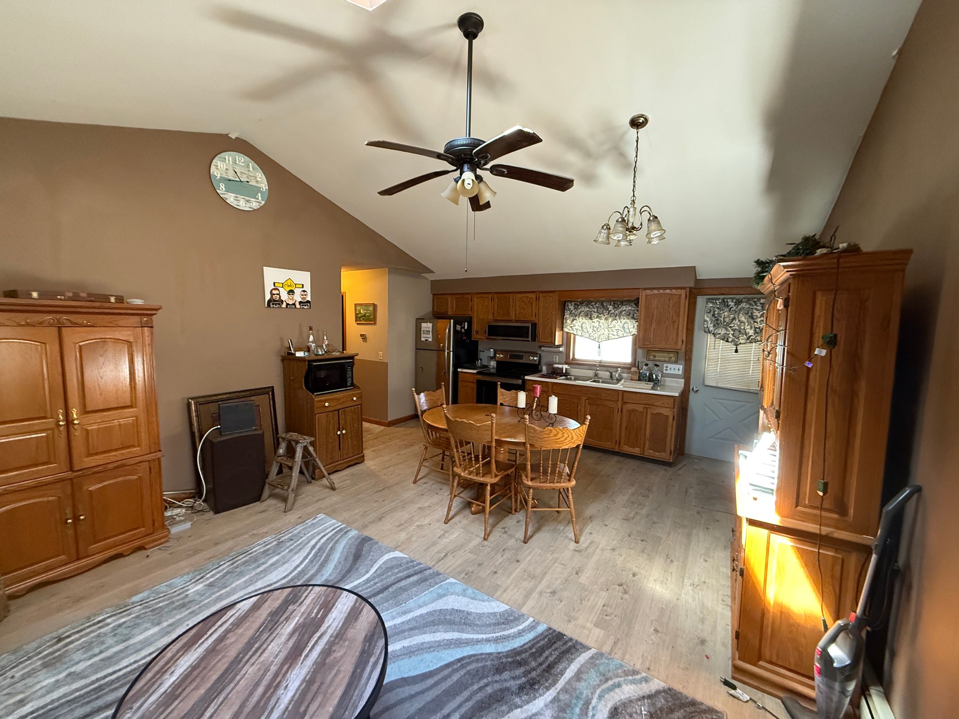 Living room with kitchen in background; brown walls and cabinets, wooden furniture, and light-colored flooring.