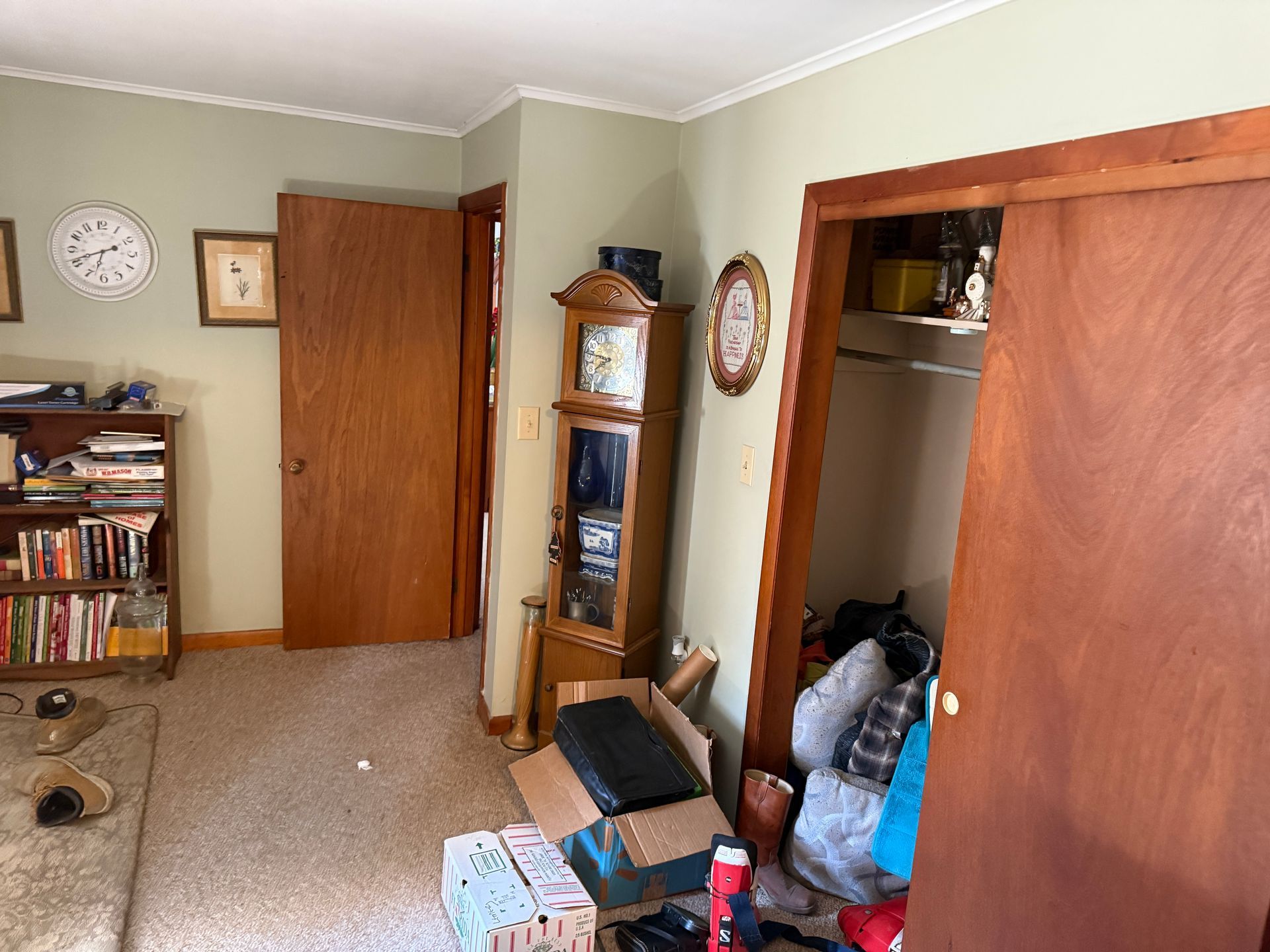 Room with tan carpet, wooden doors, a grandfather clock, and a bookshelf filled with books.