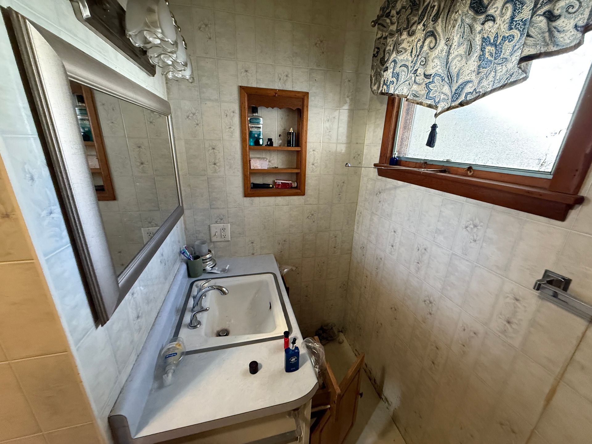 Bathroom with white tiled walls, sink, mirror, and medicine cabinet; a small window with patterned curtain.