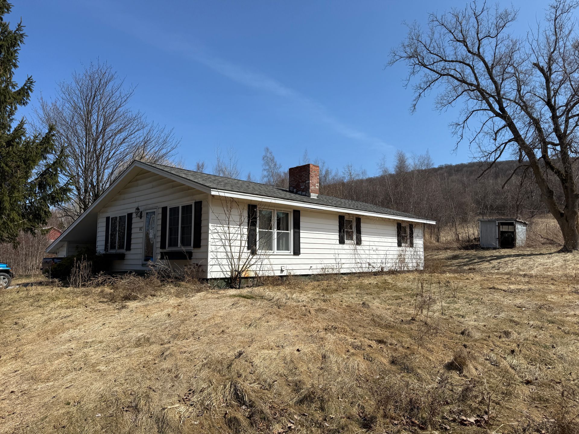 White ranch house with black shutters, brick chimney, and yard under a blue sky.