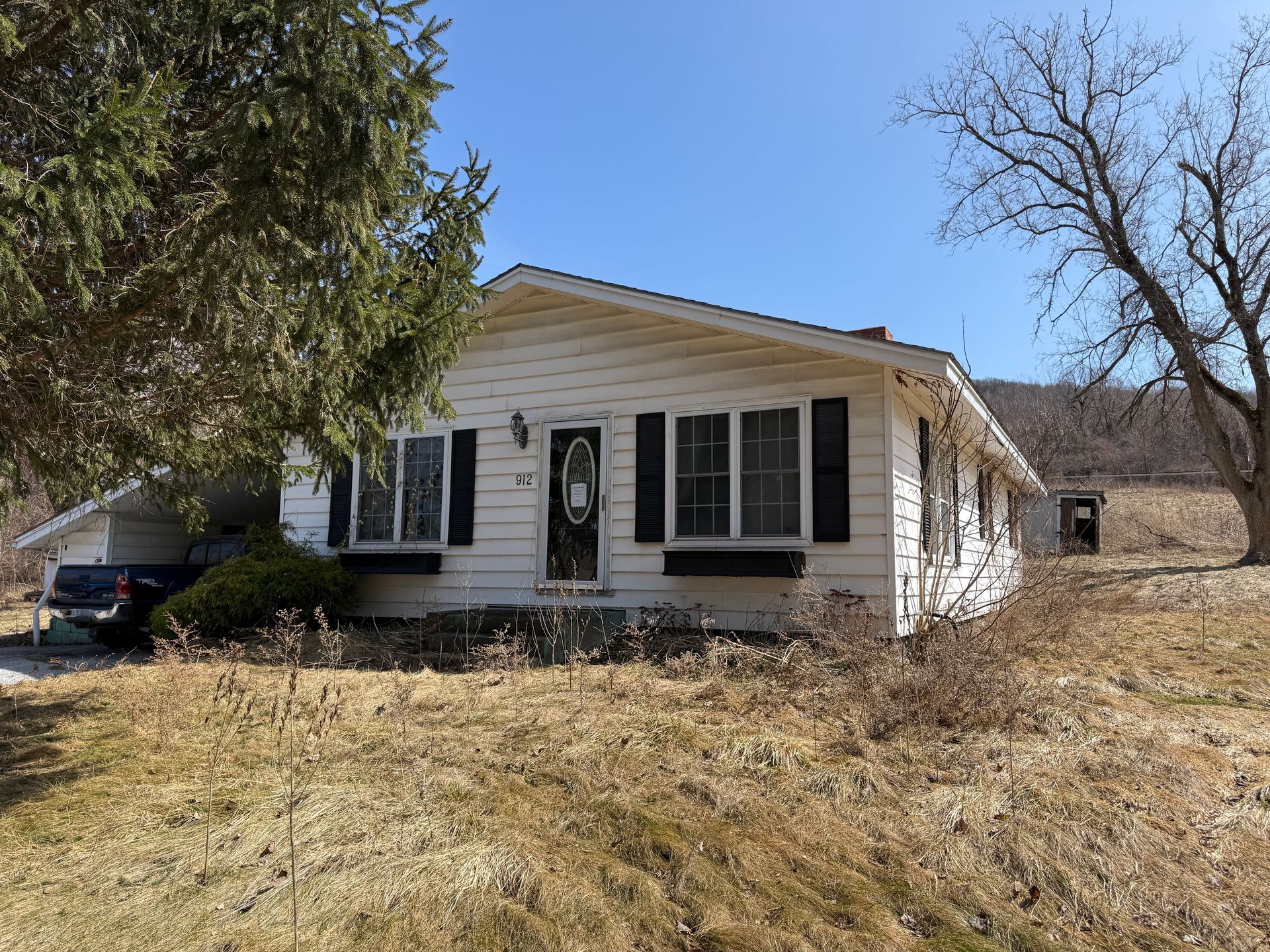 White, single-story house with black shutters, surrounded by grass and trees, under a blue sky.