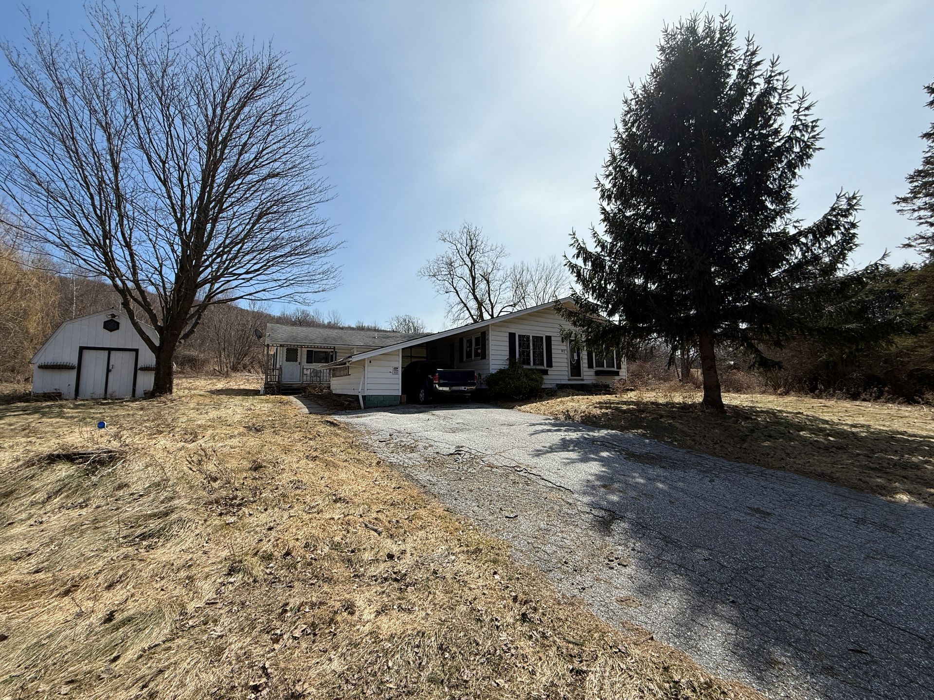 A ranch-style house with a gravel driveway and detached shed; trees surround the property on a sunny day.