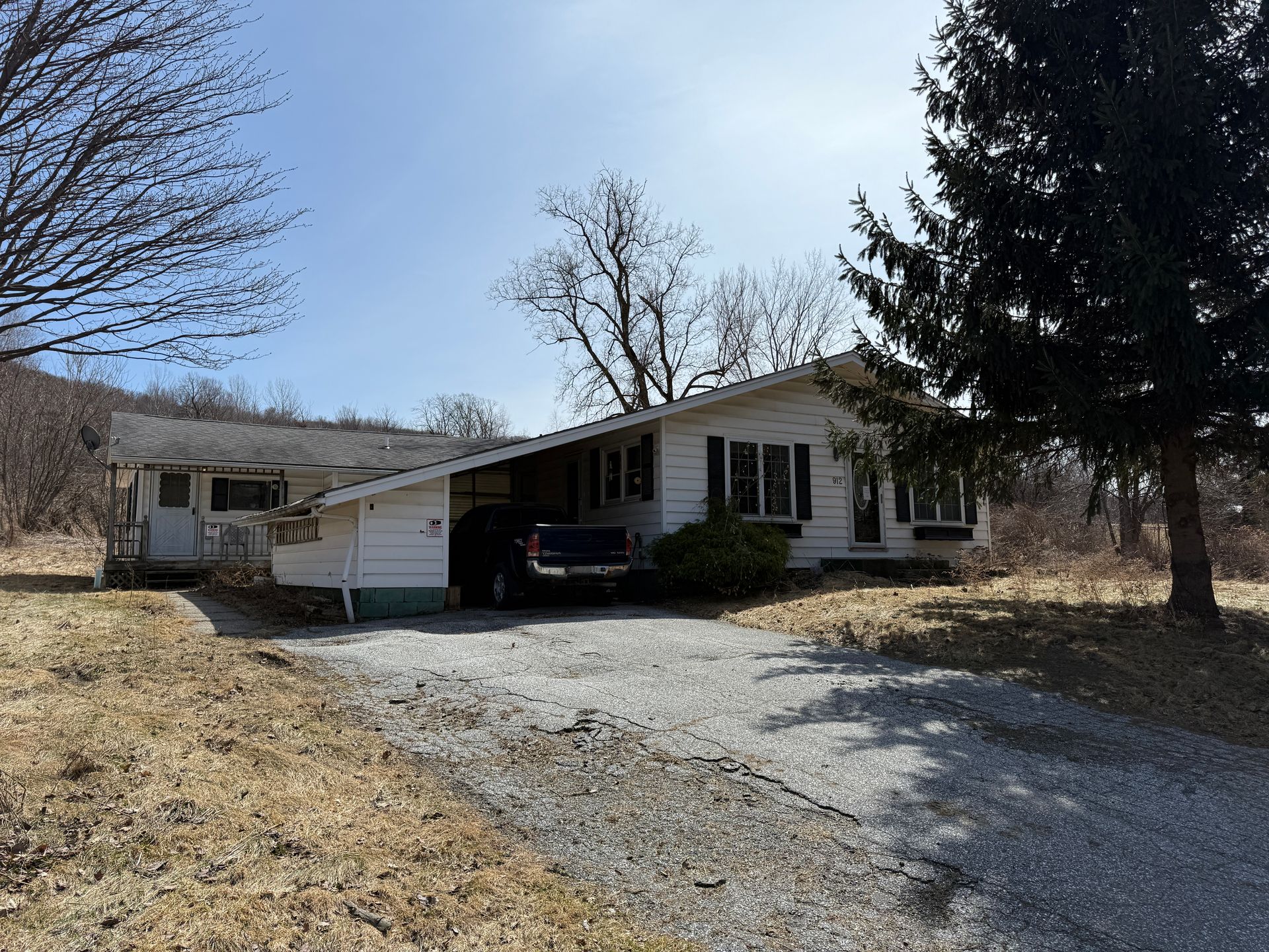 White ranch-style house with a gravel driveway, mature trees, and a parked vehicle in the garage.