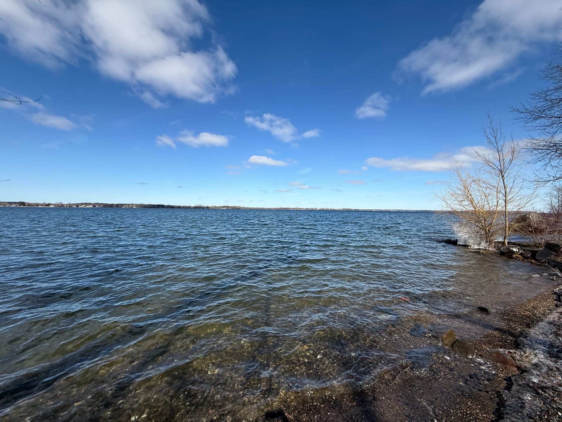 Blue lake under a bright blue sky with scattered clouds. Shoreline in the foreground.