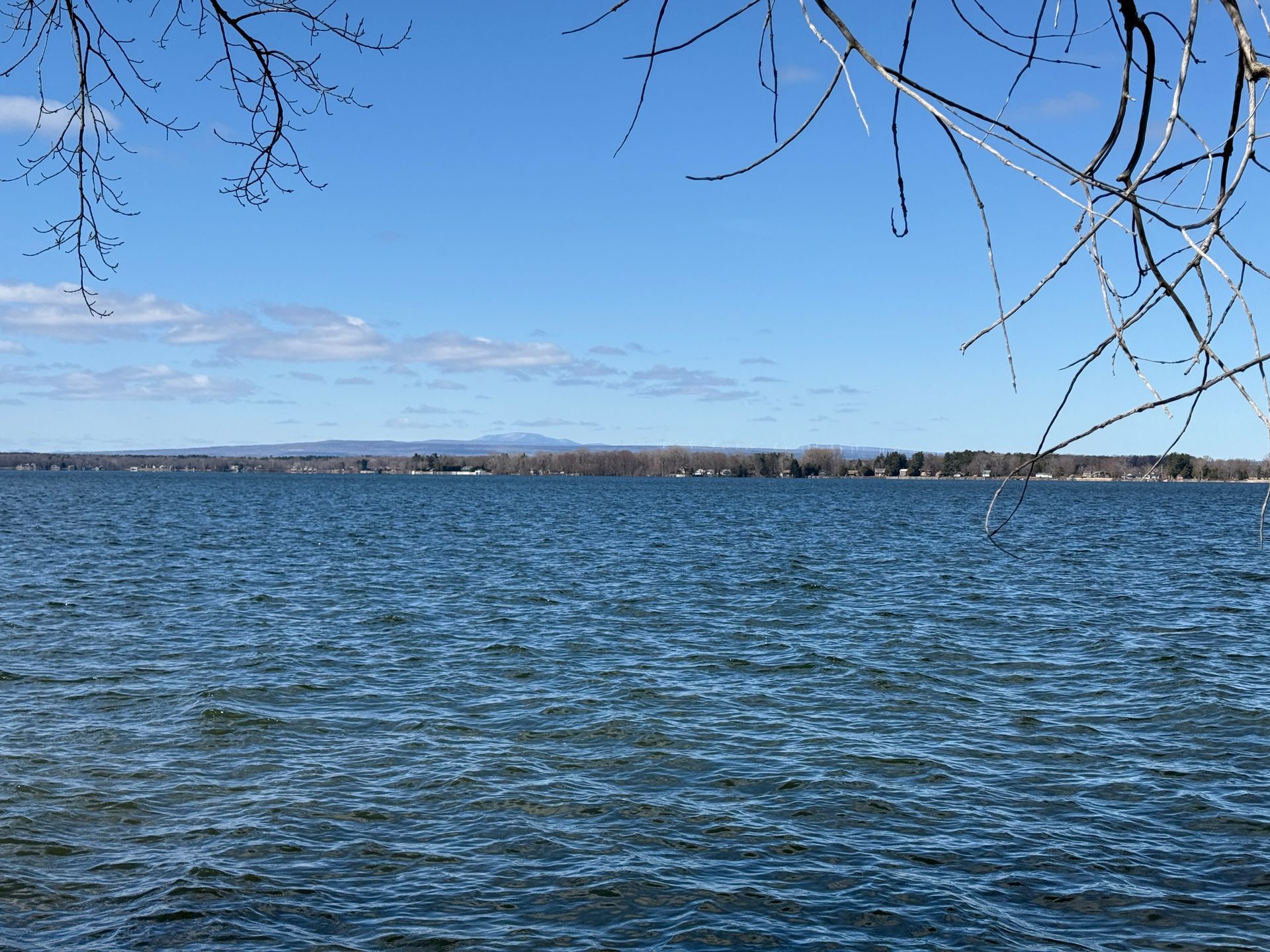 Blue lake under a clear, bright sky. Bare tree branches frame the top. Distant shoreline.