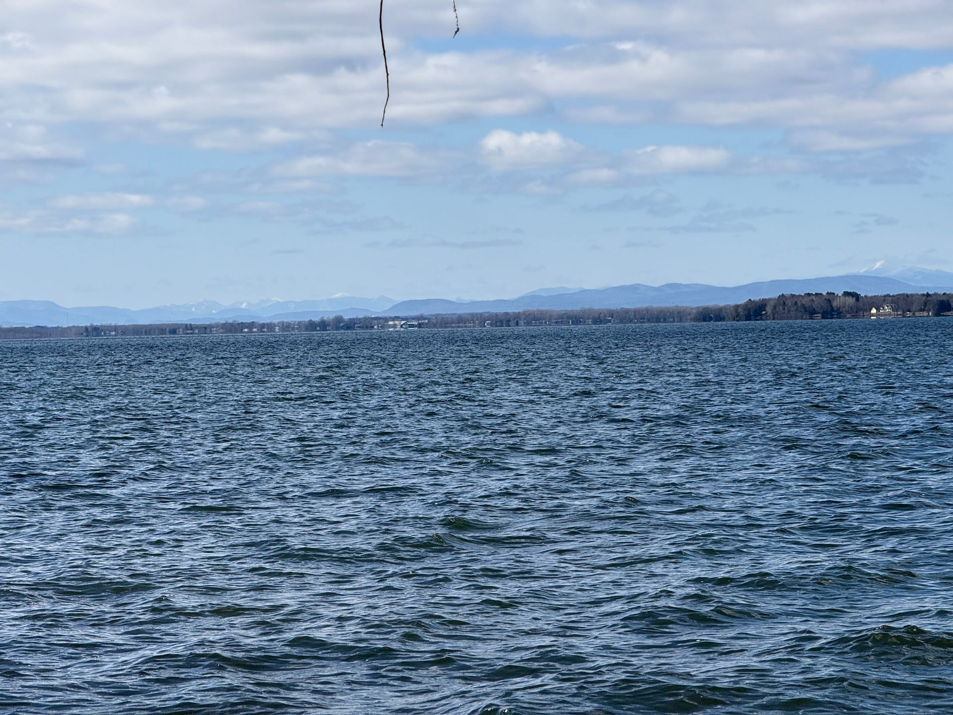 Blue water with small waves under a cloudy sky. Distant shore with mountains visible.