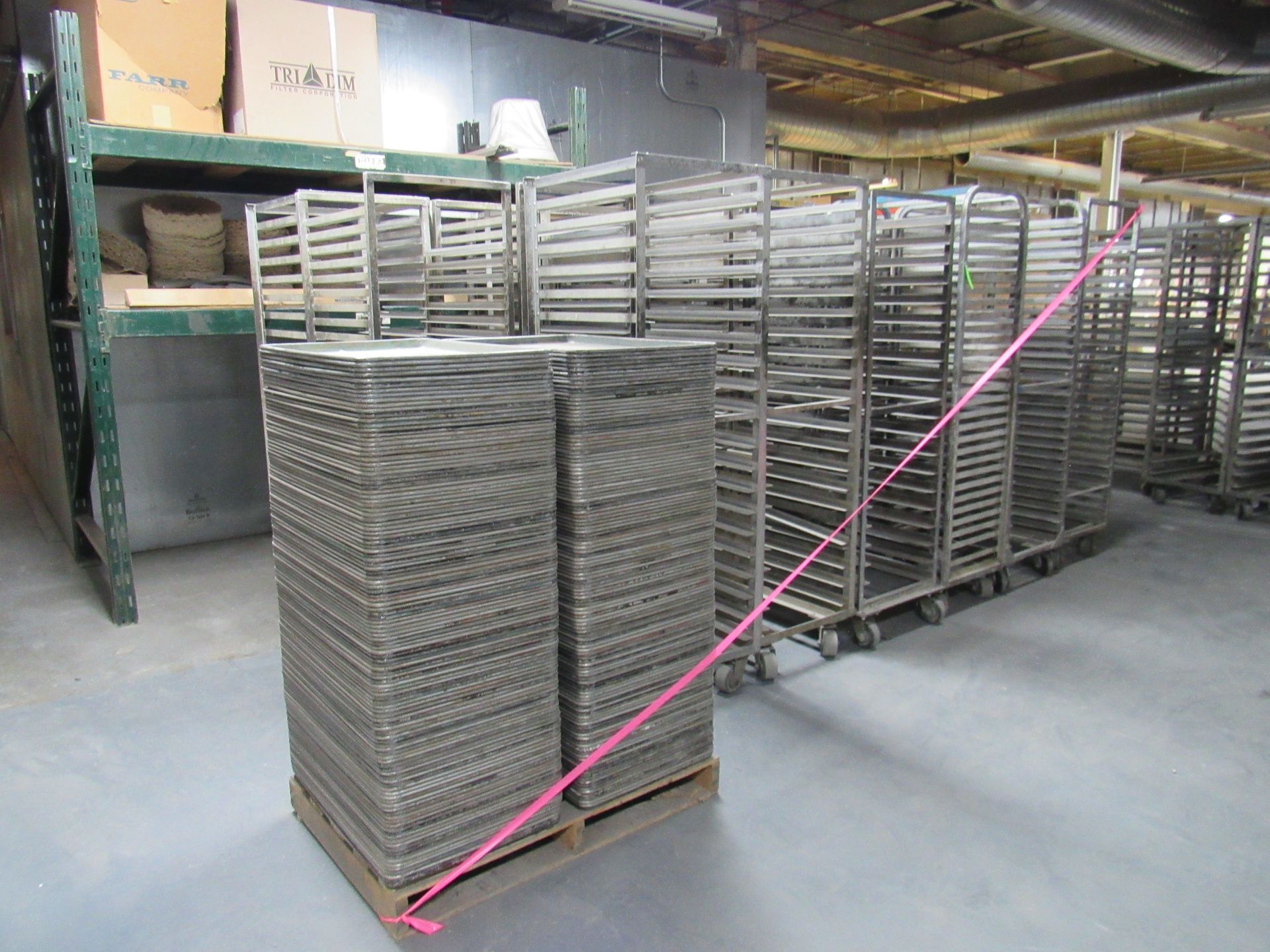 Stacks of metal trays on a pallet, beside rolling racks of trays in a warehouse setting.