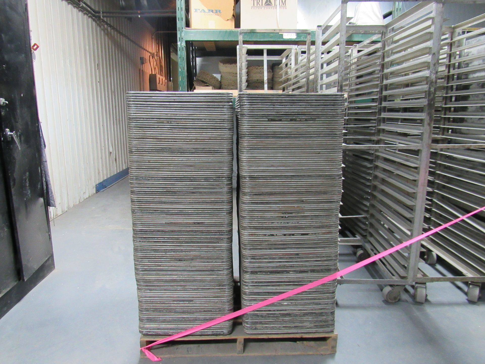 Two stacks of grey trays on a pallet, secured with pink tape, in a warehouse setting.