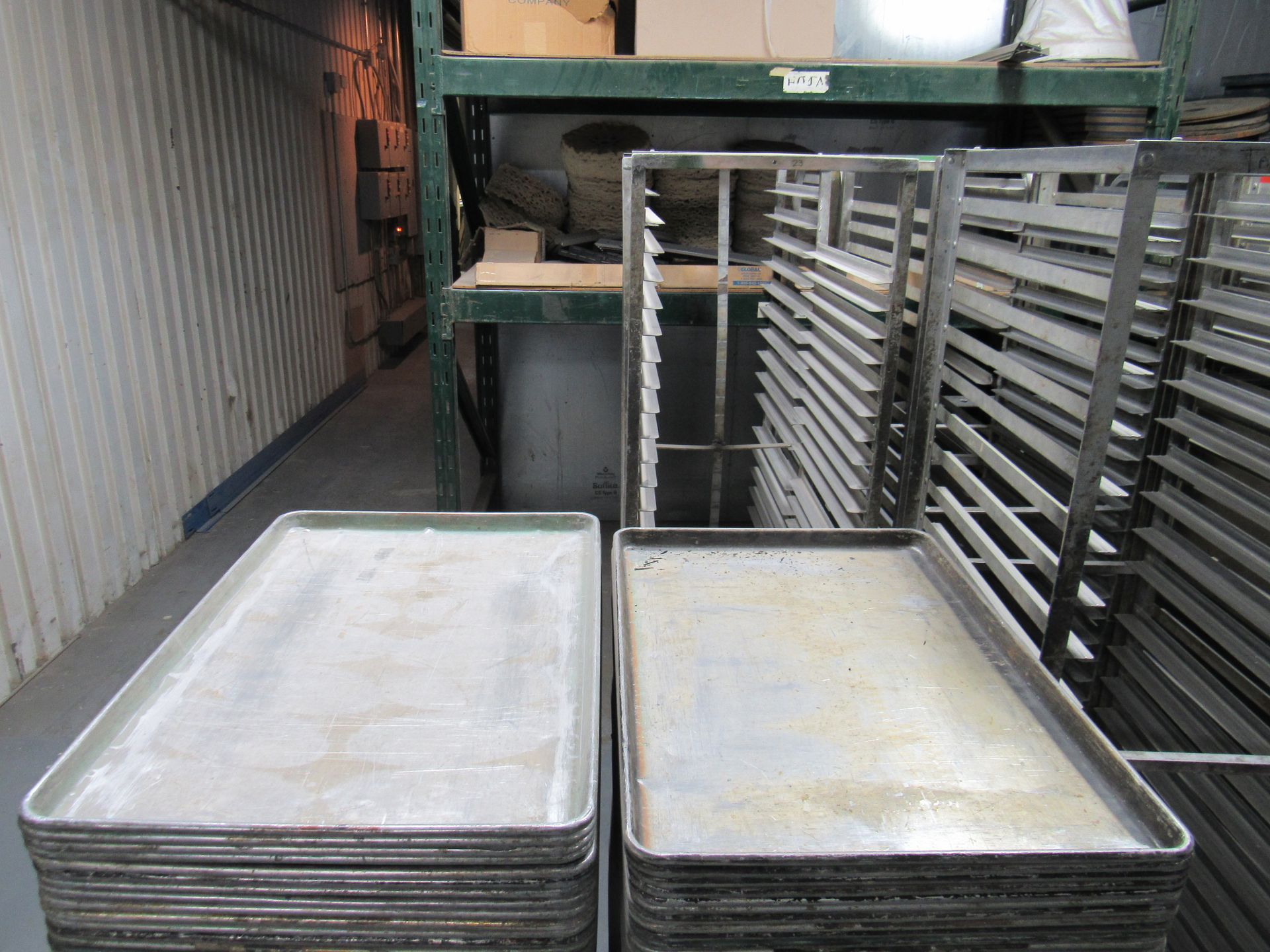 Stacks of metal baking trays in a commercial kitchen setting, with shelving in the background.