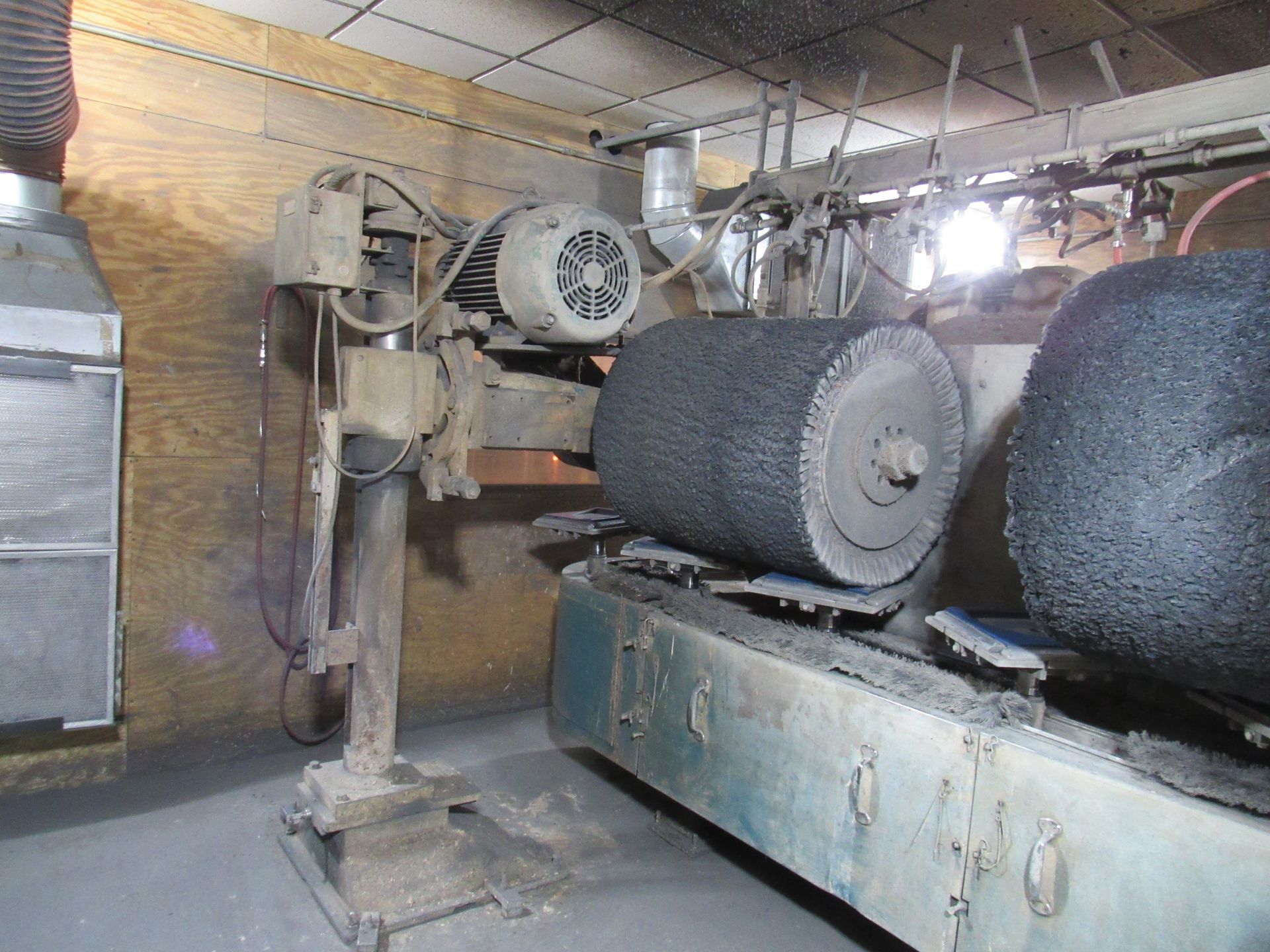 Industrial machine with two large, rotating black brush rollers; wooden panel wall in background.