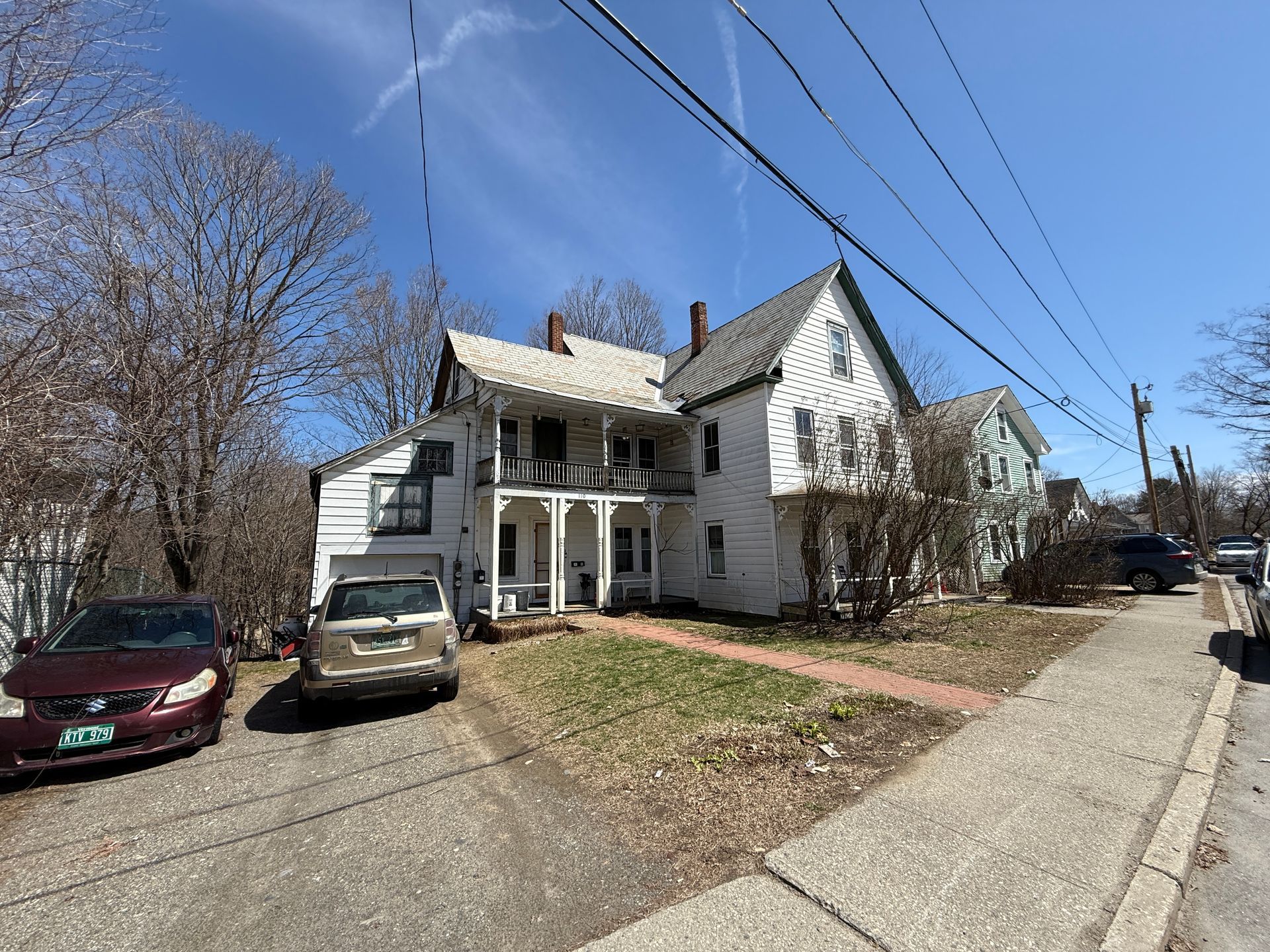 White multi-story house with porch; parked cars and sidewalk in foreground. Blue sky.