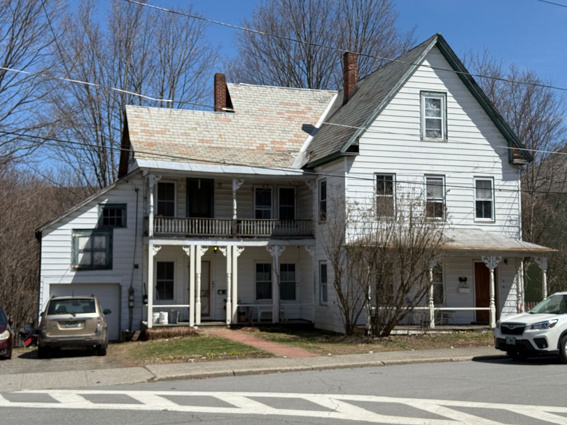 White two-story house with porch and balcony. Cars parked in front on sunny day.