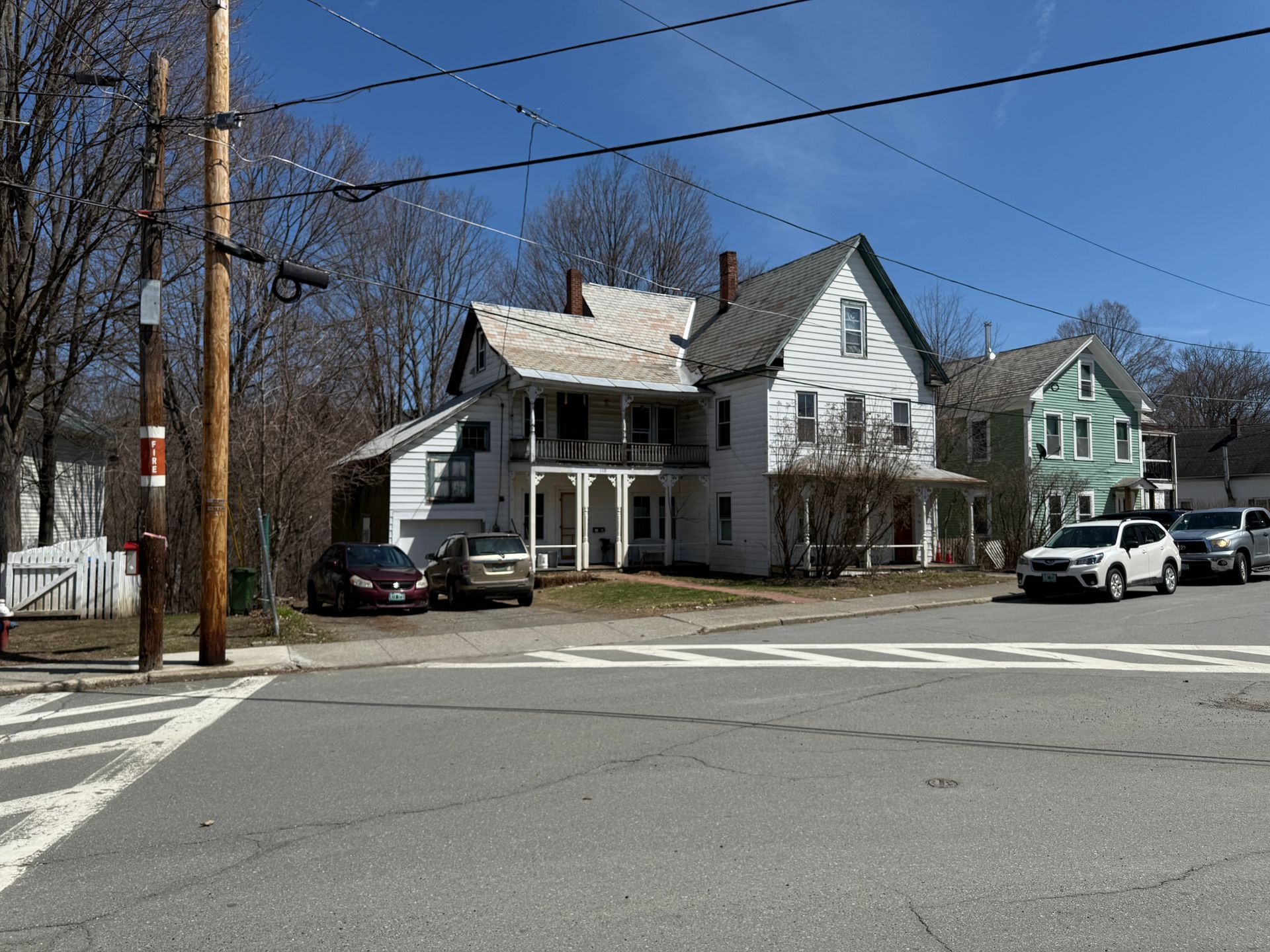 White house with porch at an intersection; cars parked, blue sky.