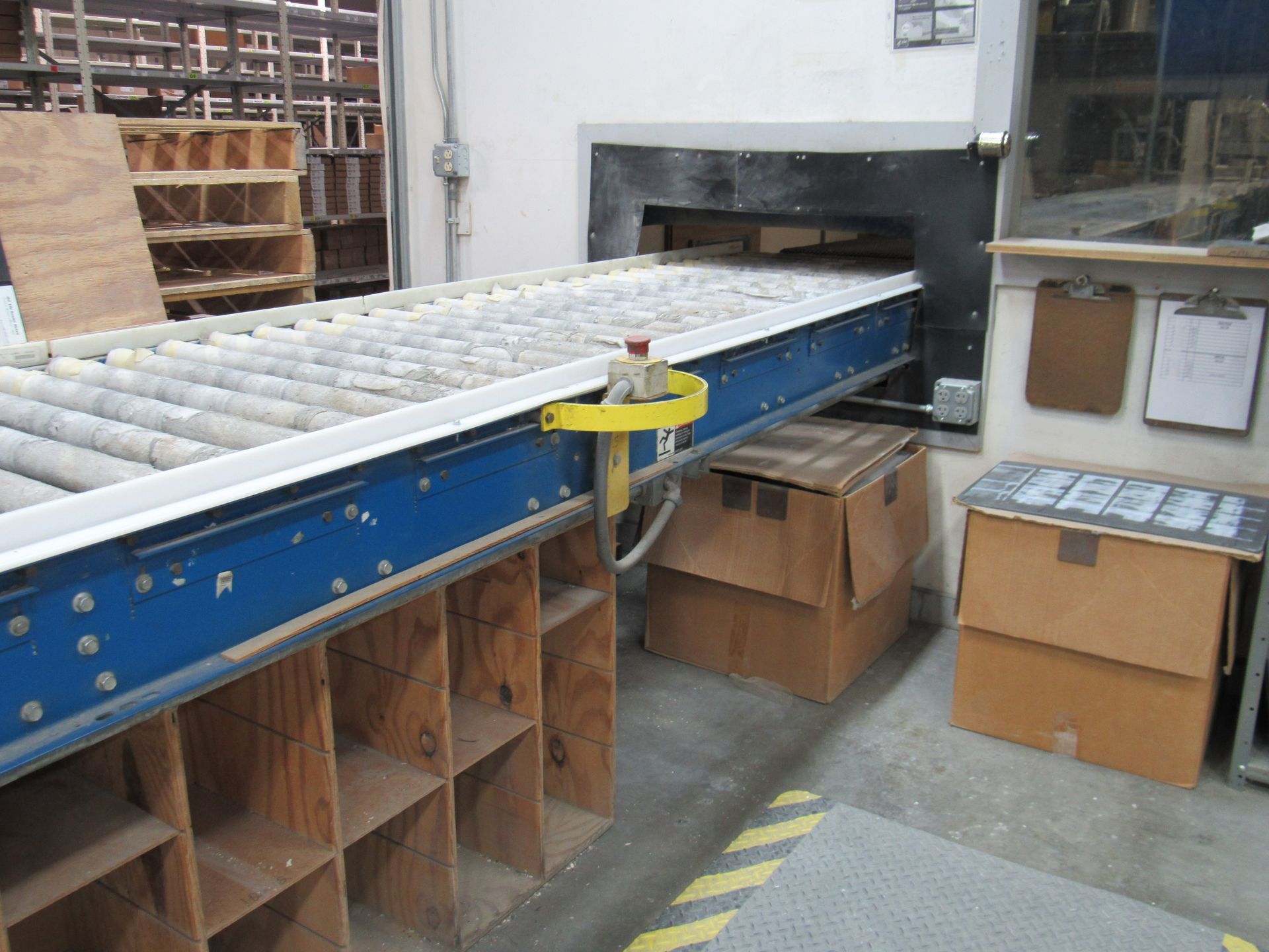 Blue roller conveyor entering a dark oven. Cardboard boxes and storage shelves nearby.