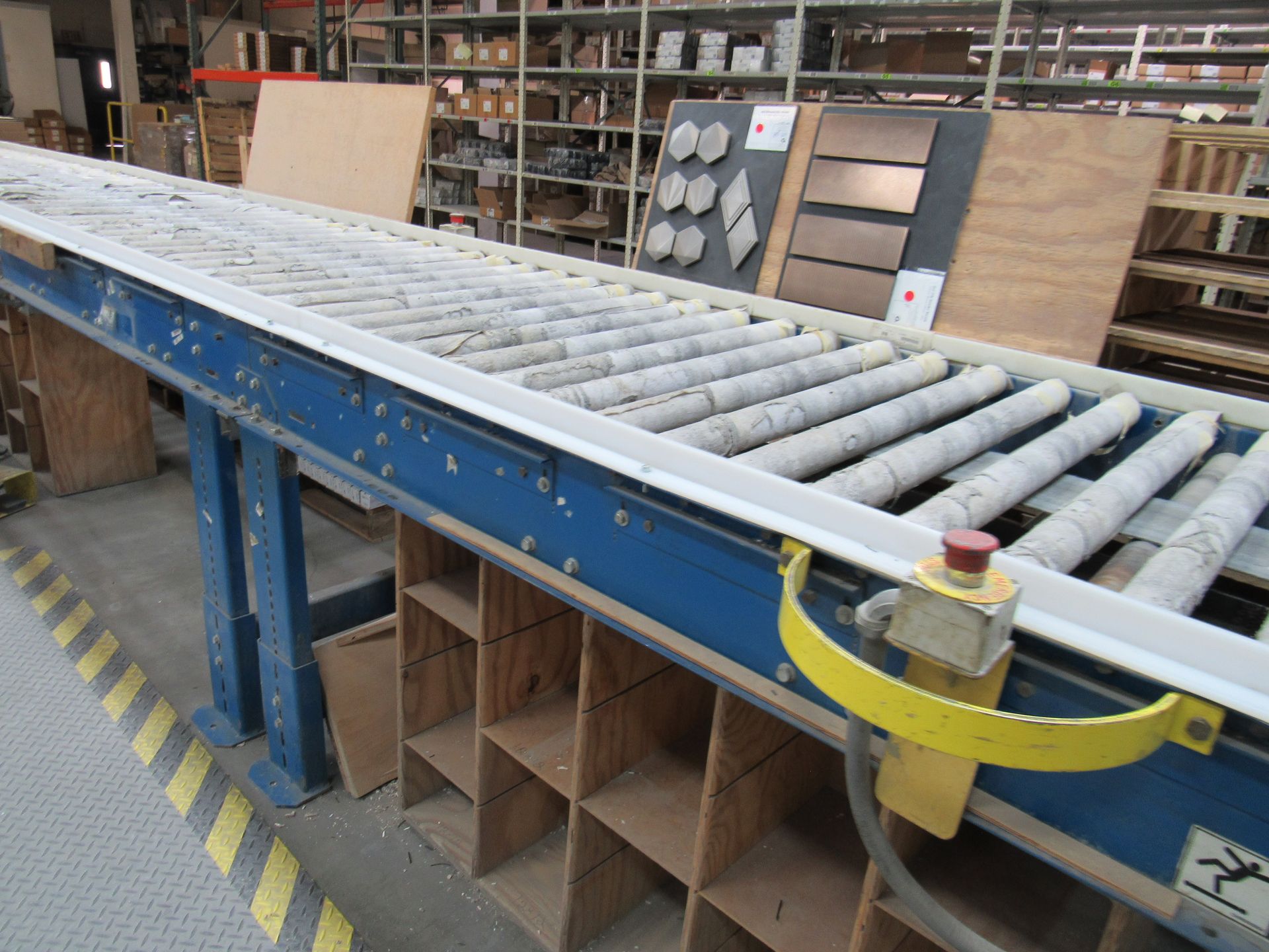 Conveyor belt with gray rollers in a warehouse, likely for moving materials. Wooden shelves in the background.