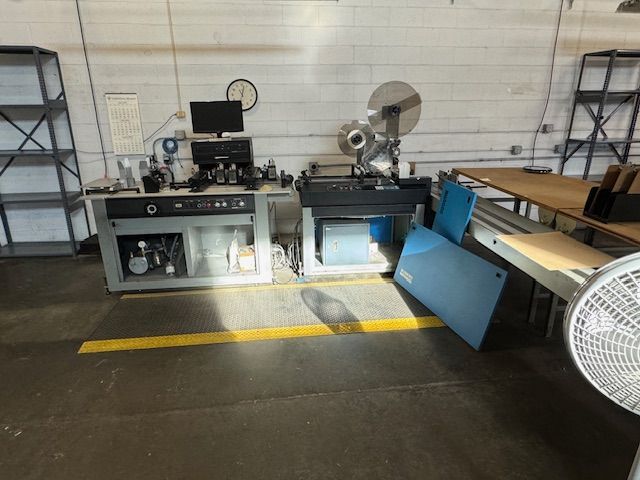 Industrial machines in a workshop: grey cabinet, printing press, fan, shelves, and a yellow floor line.