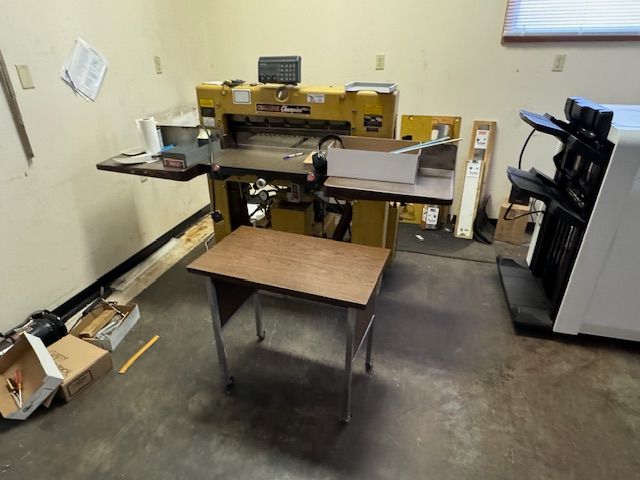 Large paper cutter machine in a room with a small desk and other office equipment.