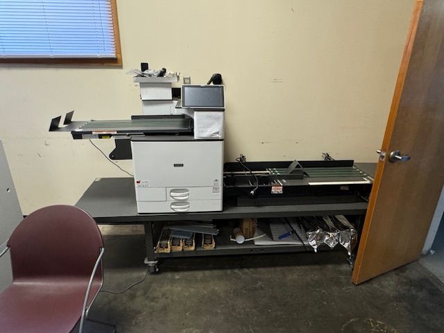 Large office printer with a feeder tray on a table, next to a door and a chair.