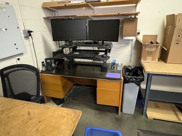 Workspace with dual monitors, desk, chair, and storage shelves in a workshop setting.