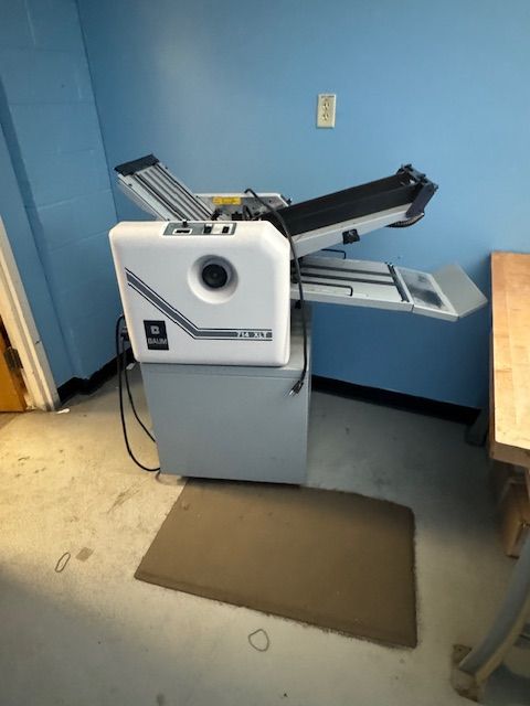 A white and gray folding machine on a gray cabinet, against a blue wall.