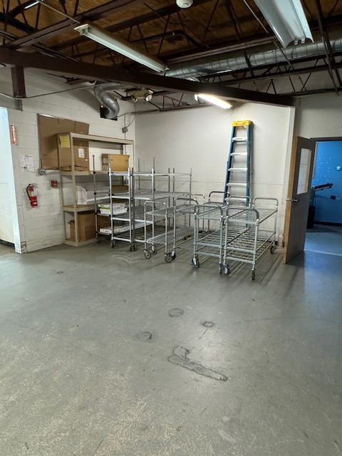 Empty industrial storage space with metal carts, shelving, and a ladder against a white wall.