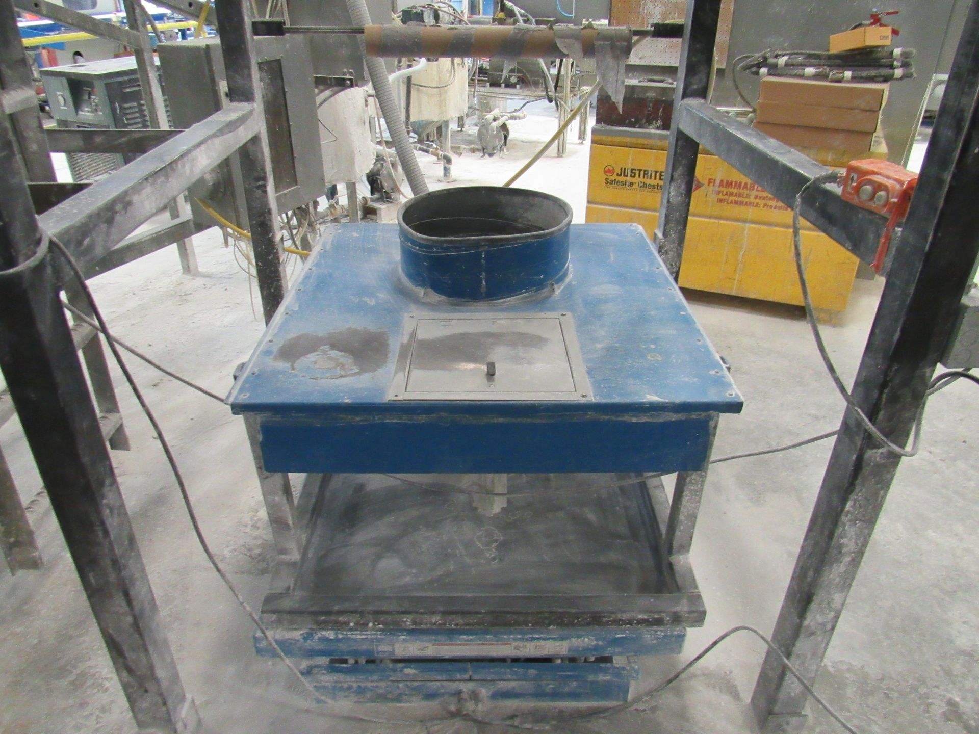 Blue table-like machine in an industrial setting. It has a round opening at the top and a tray below.