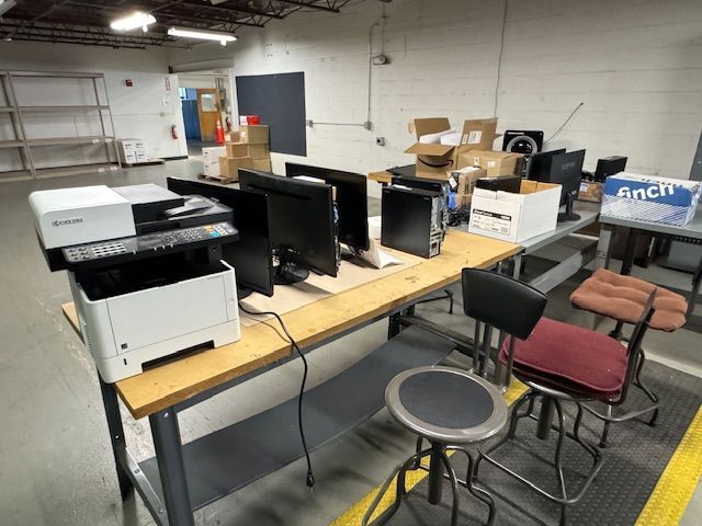A workstation with monitors, computers, printer, and stools in a warehouse setting.