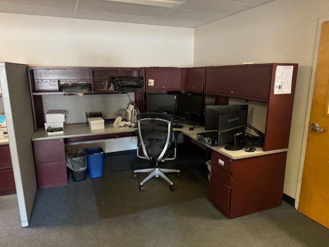 L-shaped office desk with a black office chair, two computer monitors, and overhead storage cabinets.