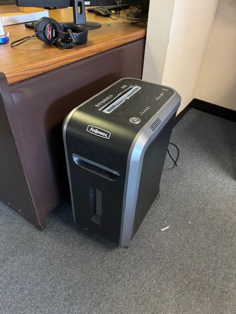 Black and silver Fellowes paper shredder on a gray carpet next to a brown desk.