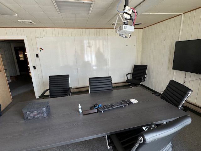 Meeting room with a large table, chairs, whiteboard, and a wall-mounted TV. Projector hangs from the ceiling.