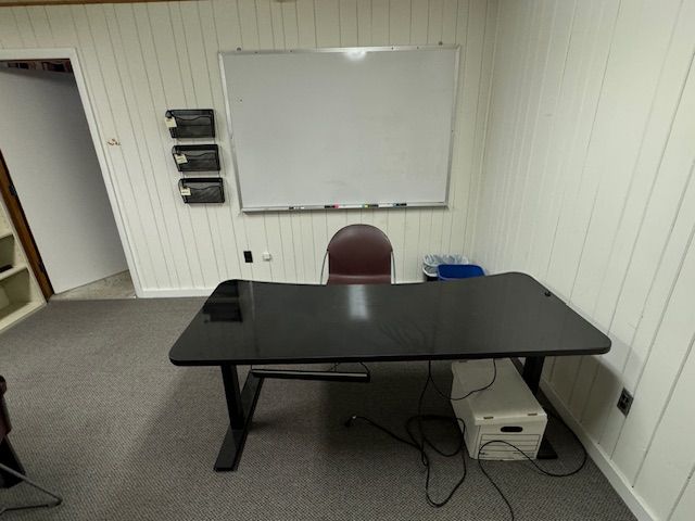 A home office with a black desk, chair, whiteboard, and printer against a white-paneled wall.