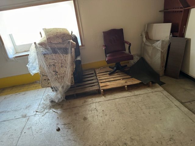 A room with pallets, an office chair, and boxes near a window.