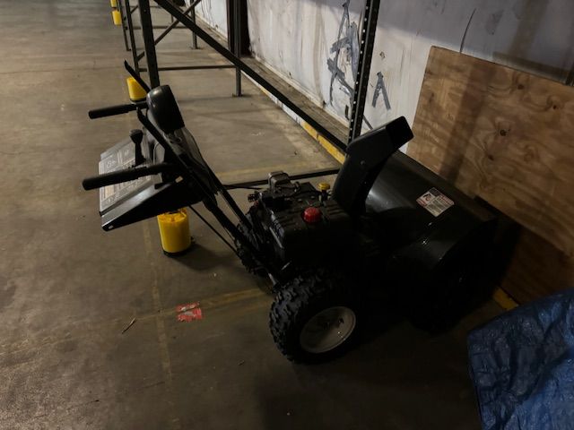 Black snowblower in an indoor setting, near a wall and shelving.