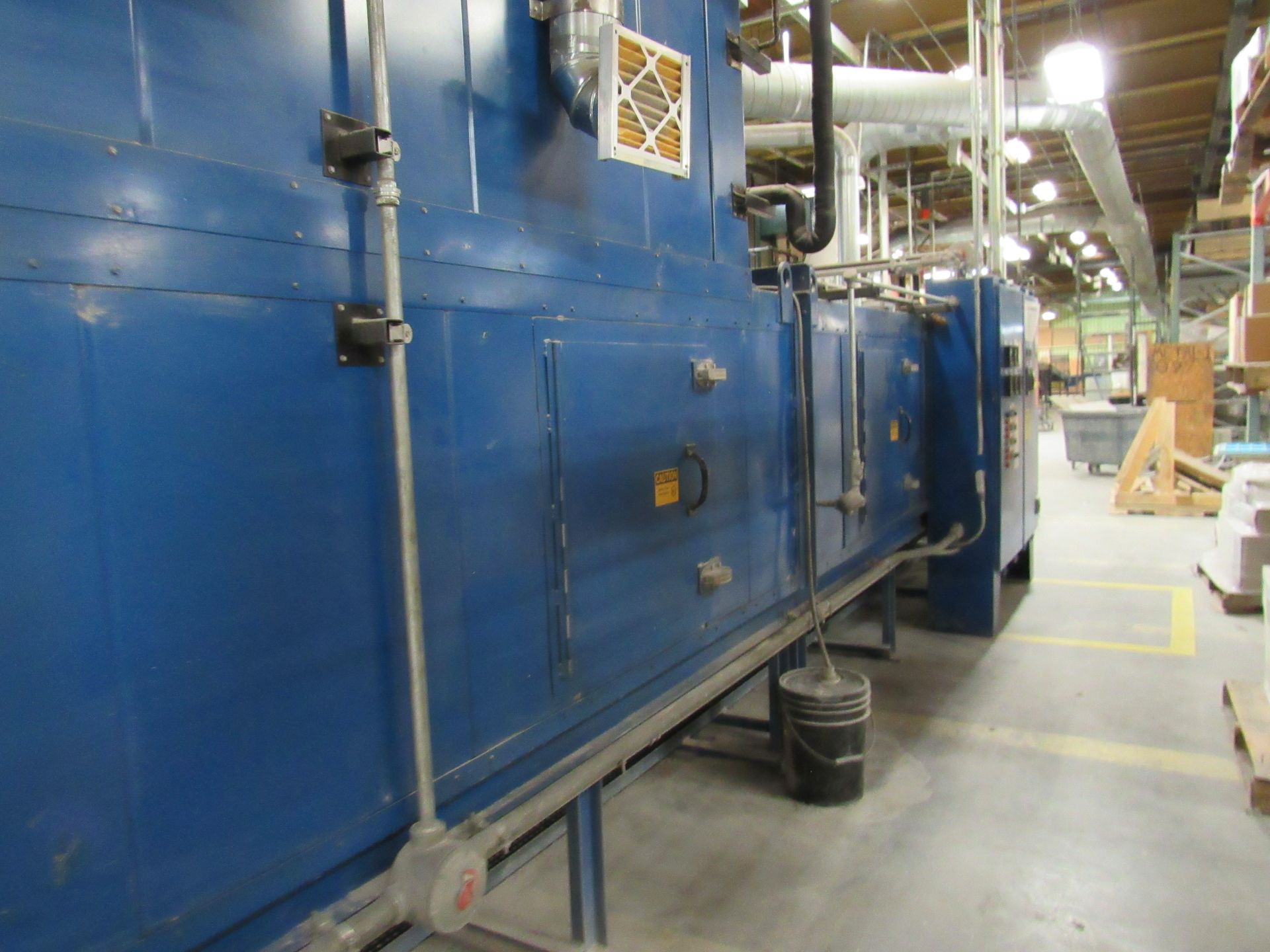 Blue industrial machine in a factory setting, with pipes and vents, near a yellow line on the floor.