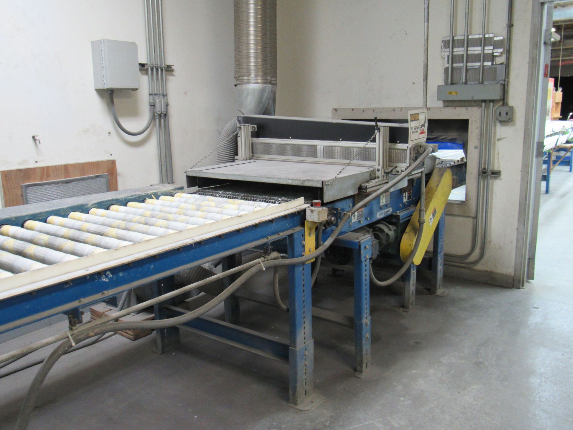 Conveyor belt with rollers leading into industrial machine with blue and yellow accents in a workshop.