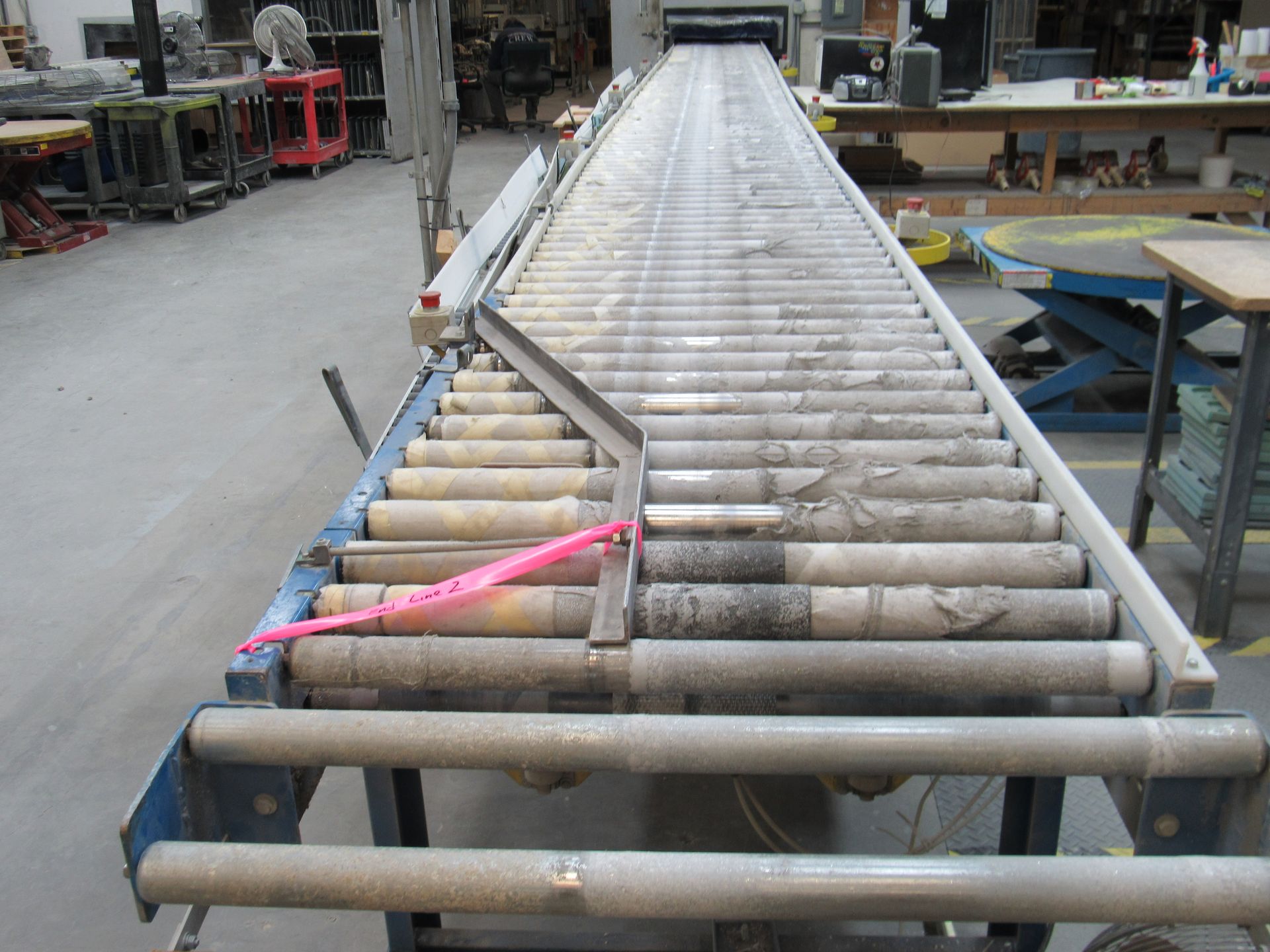 Roller conveyor in a workshop; long, horizontal with gray rollers, blue frame, and a metal ramp.