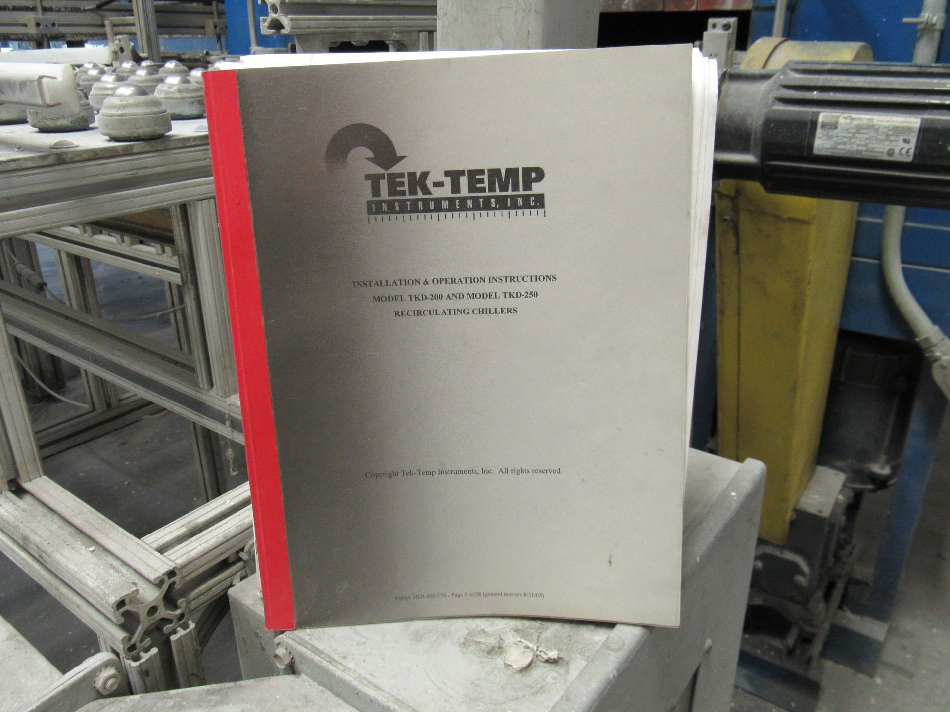 Manual for Tek-Temp industrial equipment with a red spine, on a gray surface in an industrial setting.